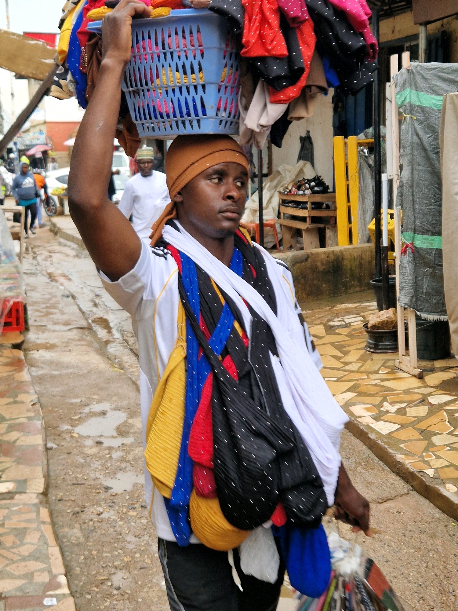 A Black male vendor is carrying a blue plastic basket filled with colorful fabrics and garments on his head. He is walking on a wet, uneven street, possibly in the city of Jos, Nigeria. The vendor is wearing a white t-shirt with black and gold stripes on the sleeves and a brown headscarf. He has several pieces of fabric draped around his neck, including yellow, blue, red, and black patterned scarves. In his right hand, he is carrying a bag filled with more textiles. The background shows a marketplace environment with buildings, market stalls, and other people. The ground is paved with interlocking stones and there are puddles of water, suggesting it has recently rained.