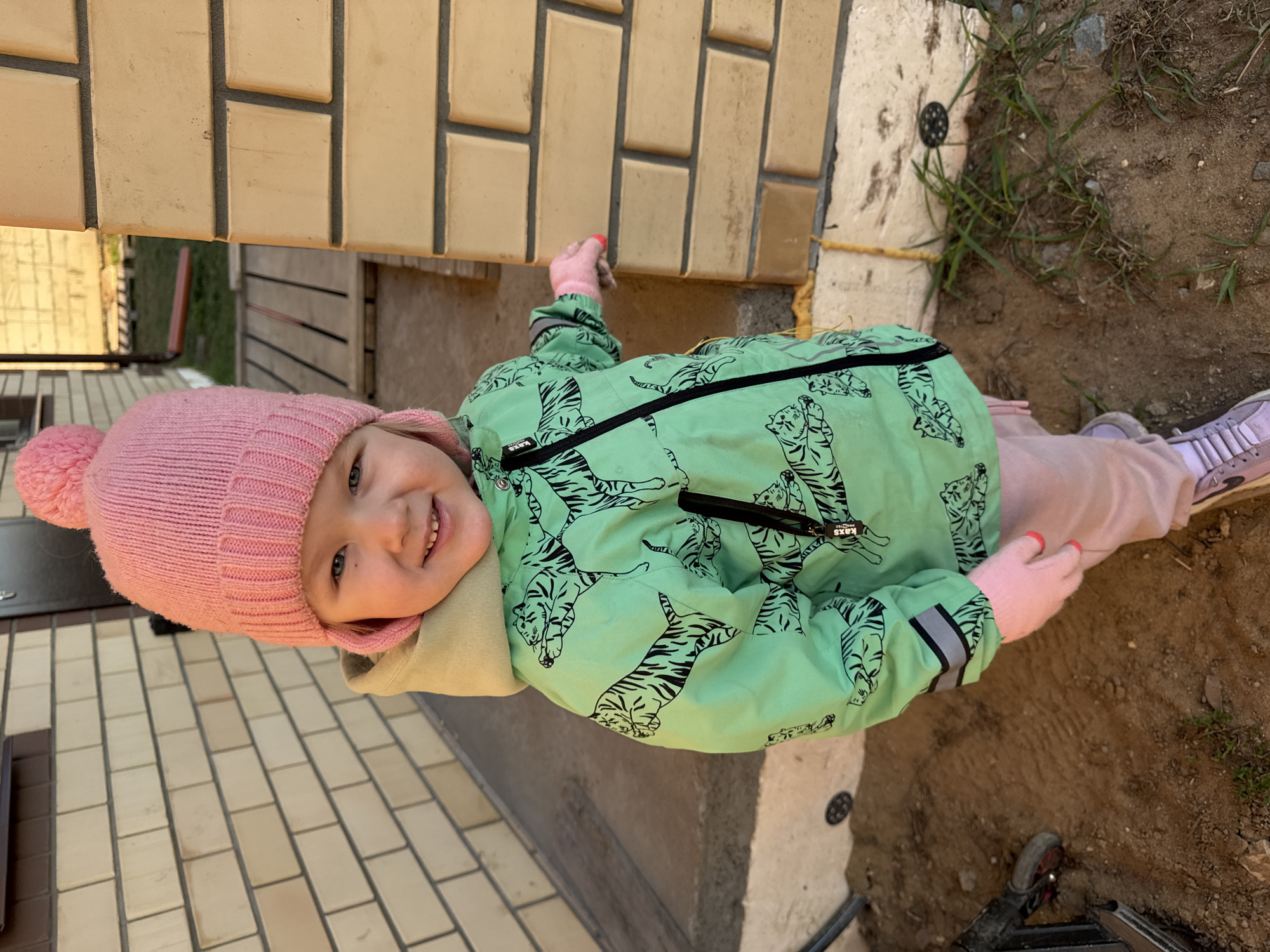 A young child, likely a toddler, stands near a building. She wears a pink knit hat with a pompom, a green jacket with tiger print, pink pants, and pink gloves. She has light skin and is smiling. The building has a tan brick exterior. Some dirt and vegetation are visible near the base of the building. This appears to be an outdoor setting. Based on location data provided in the original request, this scene is in Ферма, Russia.