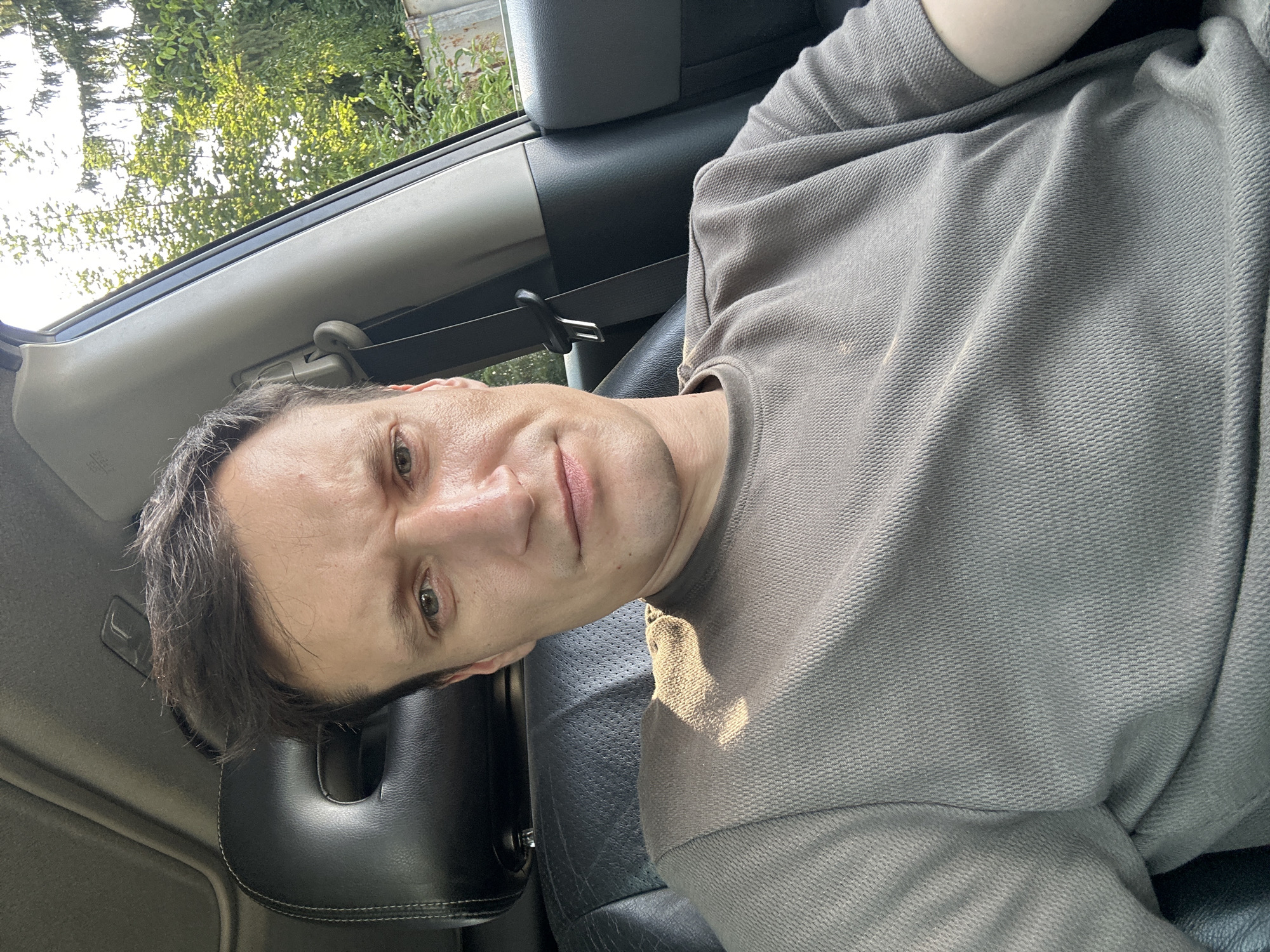 The image shows a medium shot of a man sitting in the driver's seat of a car. He is looking directly at the camera with a serious expression. The man has short, dark hair and is wearing a grey t-shirt. The interior of the car is visible, including the black leather seat and a portion of the dashboard. Outside the car window, trees and foliage are visible, suggesting a natural or suburban setting. The lighting indicates it is daytime, likely a sunny day, with sunlight casting a shadow on the man's shirt. The scene is set in Shatava, Ukraine.