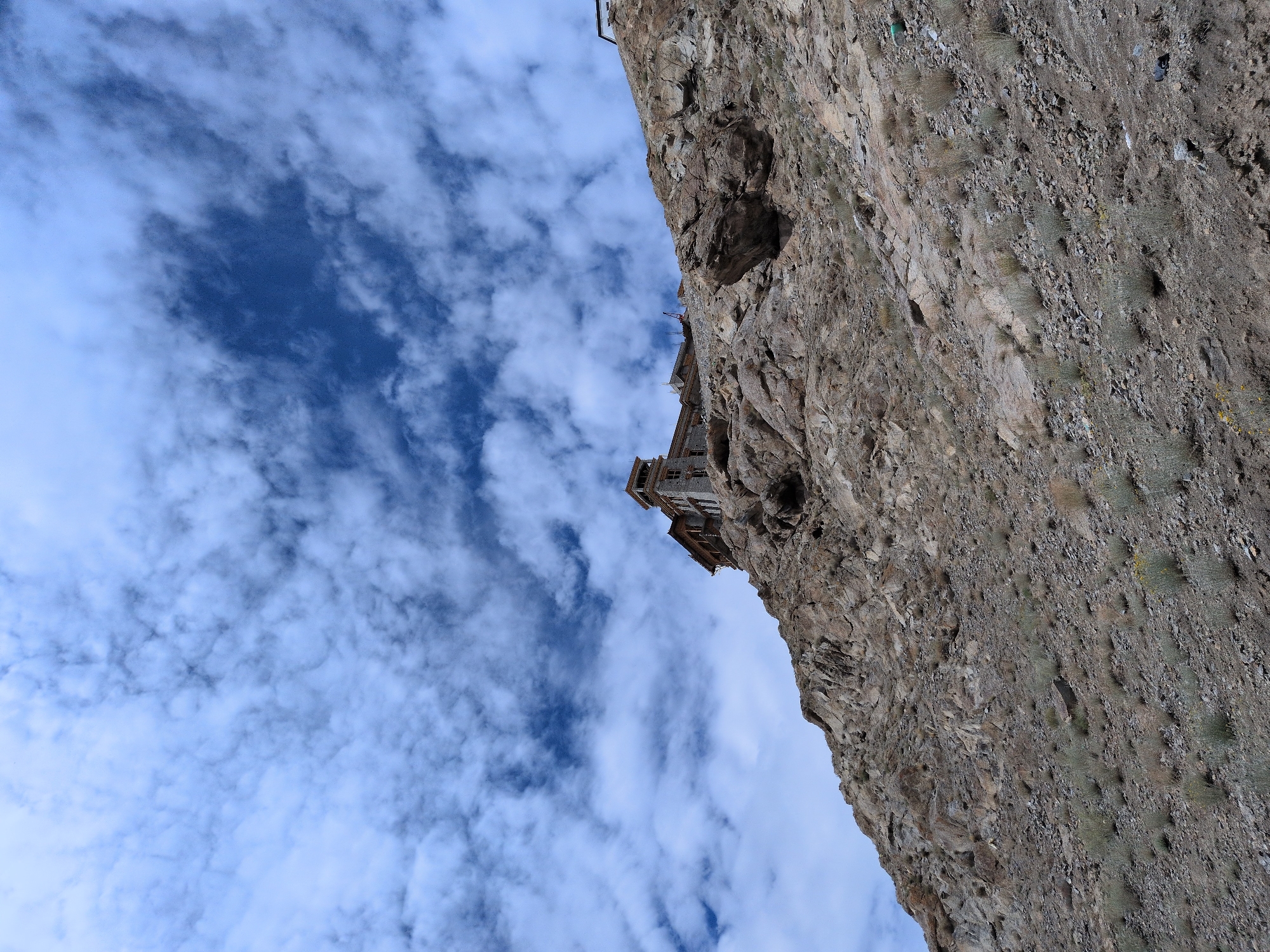 The image shows a view looking up at the Leh Palace, a historic palace and monastery located in Leh, Ladakh, India. The palace is situated on a rocky hillside with sparse vegetation. The sky above is a deep blue, dotted with white, fluffy clouds. The palace itself appears to be made of stone and has multiple levels and towers. The overall scene is one of a majestic historical structure set against a dramatic natural landscape.