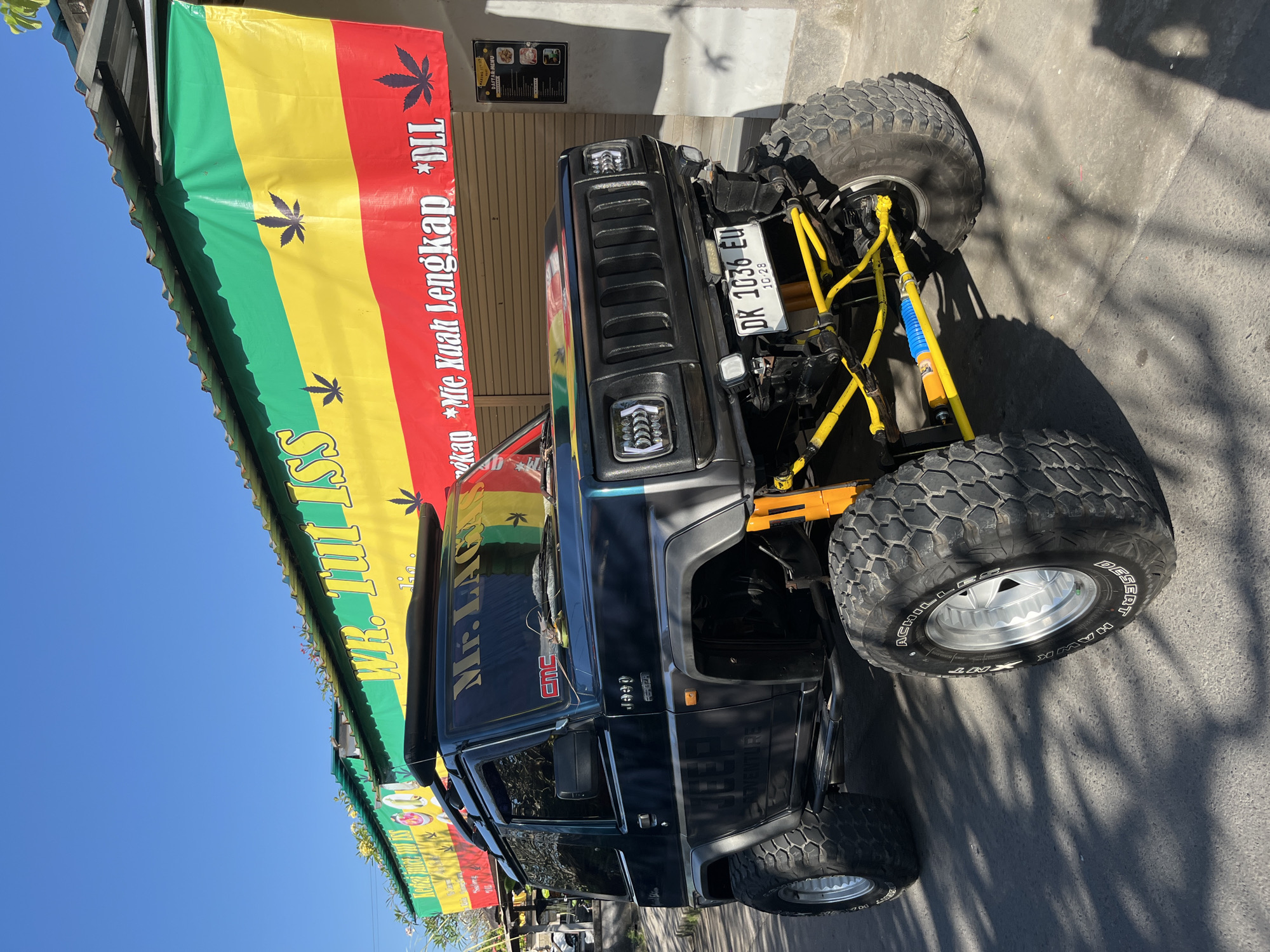 A dark green, modified Jeep is parked on a paved street in front of a building with a colorful banner. The Jeep has oversized, knobby tires and a lifted suspension system with prominent yellow components. The banner above the building is horizontally striped in green, yellow, and red, with black marijuana leaf symbols printed on it. Text on the banner reads "WR. Tui ISS" and "*Mie Kuah Lengkap *DLL". The building's facade consists of closed brown shutters. A menu board is attached to the right wall of the building. The sky is clear blue, and shadows from the Jeep and foliage are cast on the ground. The image appears to be taken in Sukawati, Indonesia, given the license plate format "DK 1036 EU", which is typical for Bali.