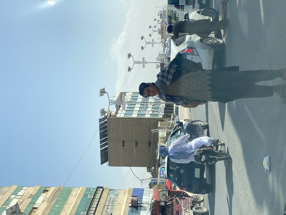 The image depicts an urban street scene in Kandahar, Afghanistan, under a clear, bright daylight sky.
In the immediate foreground, a young male stands facing the camera, slightly smiling. He is dressed in a dark skullcap, a plaid scarf draped over his shoulders, a light-colored patterned tunic, dark trousers, and sandals. A crumpled white object rests on the asphalt directly in front of him.
Mid-ground elements include a line of traffic. On the left, a red three-wheeled motorized cargo vehicle is visible. A person wearing a white shalwar kameez and a blue and white checkered head covering is seen riding a black motorcycle, positioned behind a dark blue hatchback car. To the right, a white hatchback car is present, with another individual in dark clothing bent over near its rear. Concrete barriers separate traffic lanes further to the right. Numerous streetlights, equipped with solar panels, are visible along the thoroughfare.
The background features several multi-story buildings. On the far left, a tall building with a cream-colored facade and green-tinted windows is prominent. Centrally, a brown multi-story building with flat rectangular panels and a rooftop array of solar panels is visible. To the right, another multi-story building with light blue windows and a white facade also features rooftop solar panels. Utility lines traverse the sky, which exhibits a bright sun flare in the upper right quadrant.