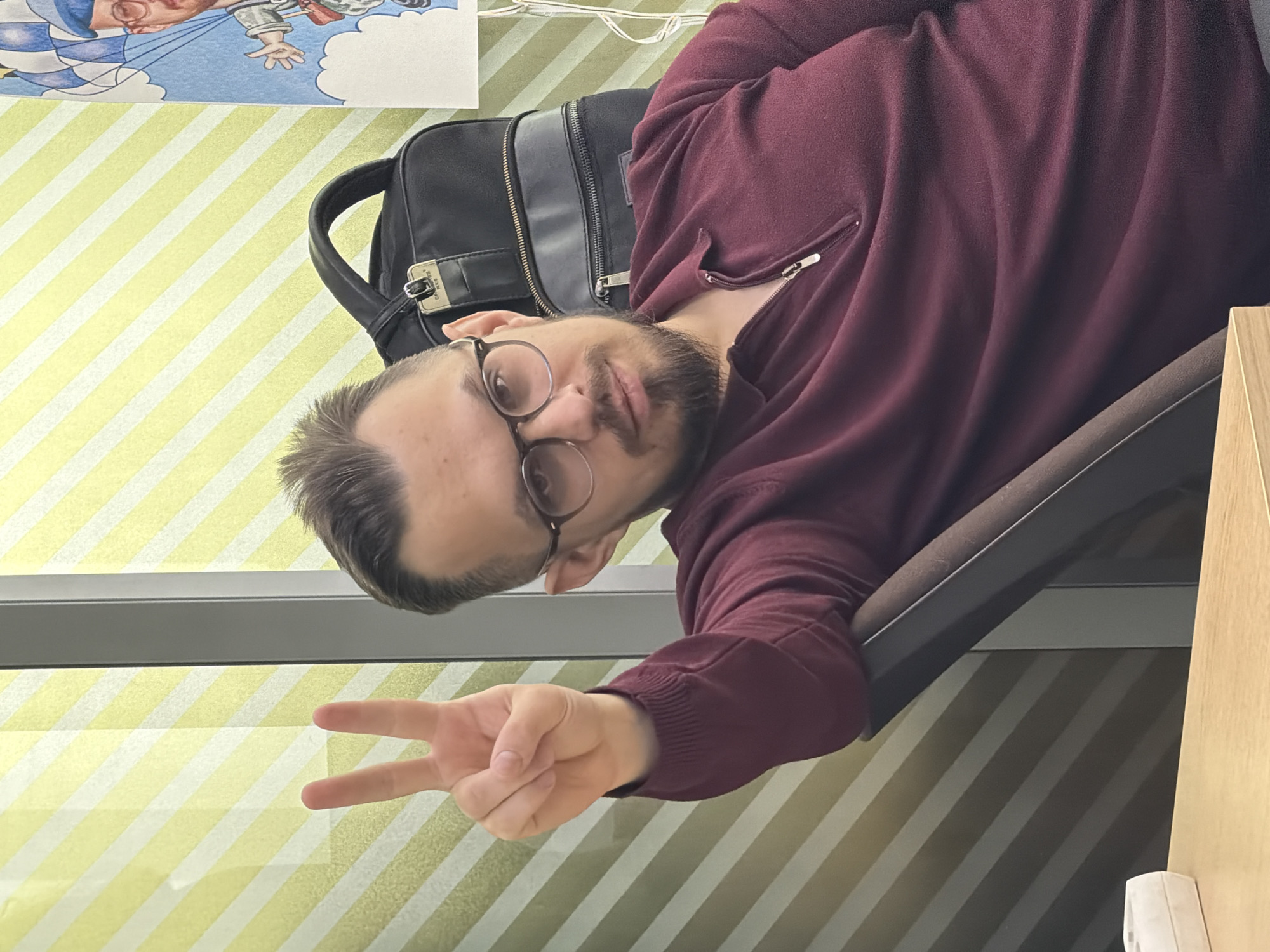 A medium shot captures a man indoors, making a peace sign with his right hand. He is wearing glasses and a maroon zip-up sweater. A black backpack rests behind him, and a cartoon image is visible in the upper left corner. The background features a striped wall. The location is likely an office or workspace. The city and country details, Krasnoyarsk, Russia, are provided for contextual information.