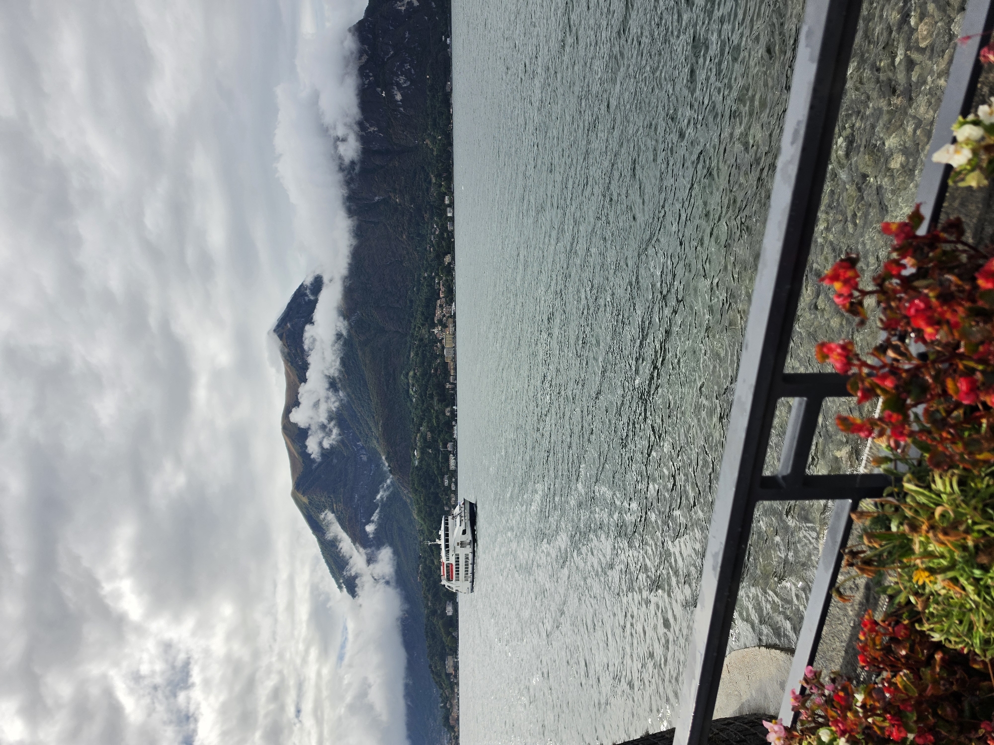 A ferry sails across a lake, with a mountain range in the background partially obscured by clouds. The water is choppy and reflects the cloudy sky. In the foreground, there is a railing and some flowering plants. The scene appears to be located in San Giovanni, Italy, though this cannot be confirmed from the image alone.