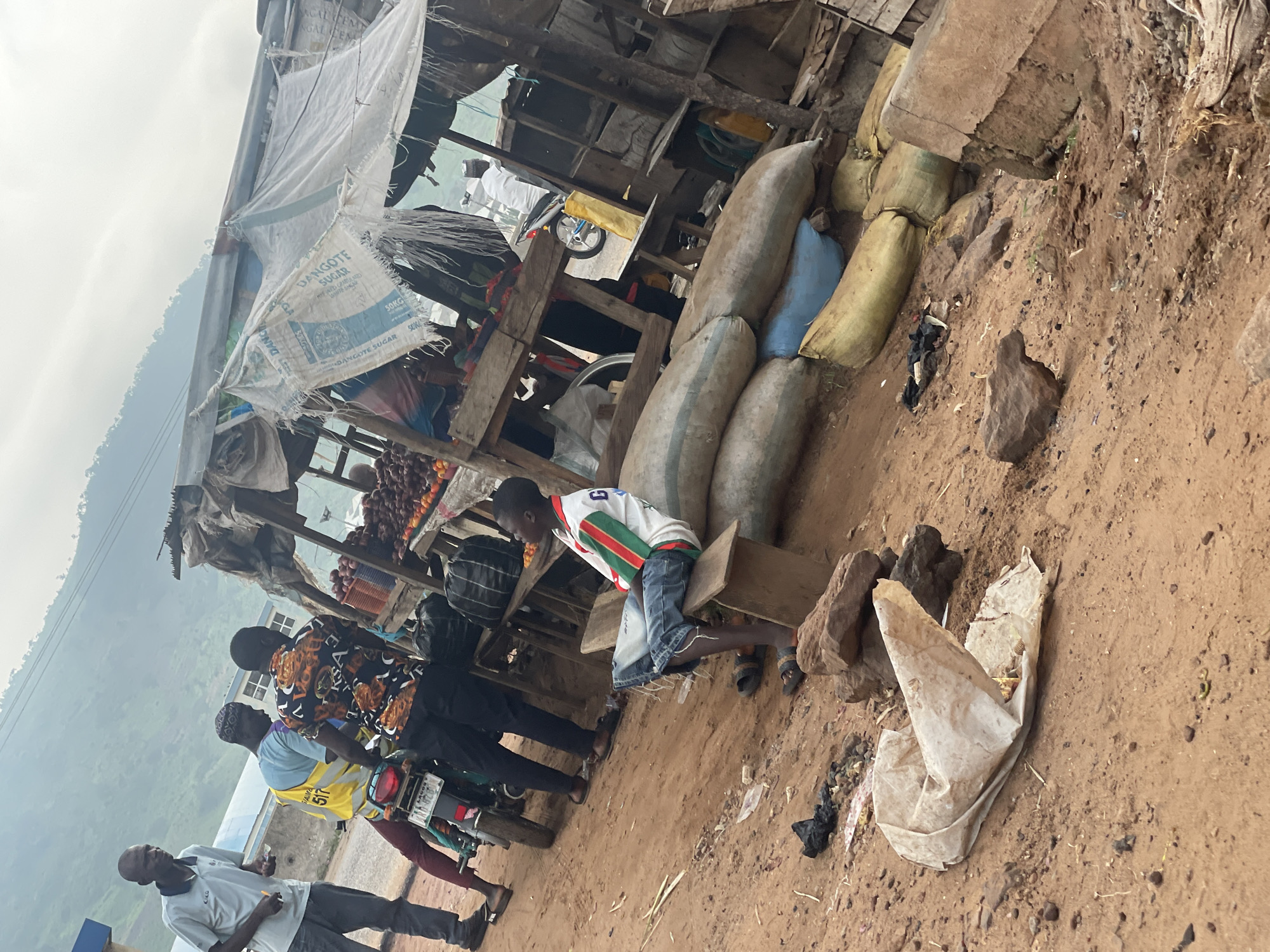 The image depicts a busy roadside market scene in Lokoja, Nigeria. In the foreground, a young boy in a white and green-striped shirt and denim shorts sits on a wooden bench. To his left, large sacks are piled up, some yellow and some blue. The ground is a dusty, reddish-brown earth, scattered with rocks and debris, including a crumpled white plastic bag and black plastic waste.
Behind the boy, a motorcycle is parked. Three men are gathered around it. One man, wearing a grey polo shirt and dark trousers, stands on the left, holding a small item. Another man, in a bright yellow vest over a blue shirt, is seated on the motorcycle. A third man, wearing a black shirt with a vibrant orange and white pattern, stands to the right of the motorcycle, facing away from the camera.
Further back, a makeshift market stall is set up with wooden structures and a tattered white tarp overhead, displaying a selection of fruits, possibly apples and tomatoes, on shelves. The stall appears weathered and rustic. Beyond the stall, a person on a motorcycle is visible, and a hazy mountainous landscape forms the backdrop under a bright, overcast sky. The overall impression is of a typical roadside market in a developing urban area, characterized by its informal structures, lively activity, and a sense of daily commerce.