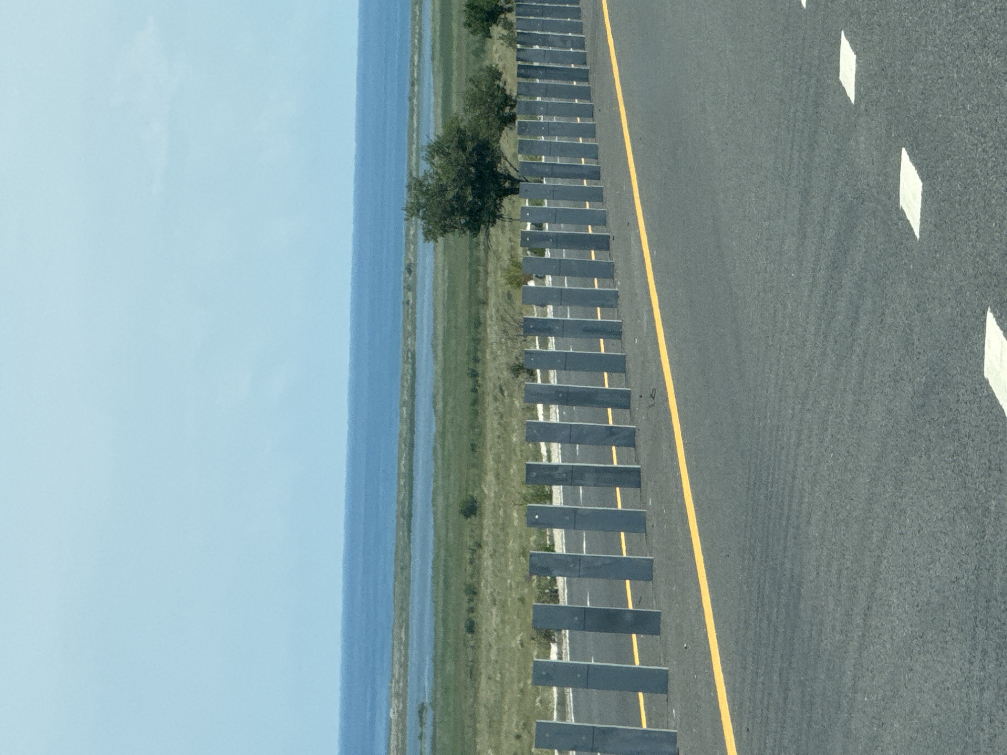 The image depicts a roadside scene with a clear blue sky and a body of water in the background. In the foreground, a grey asphalt road runs diagonally from the bottom left to the top right. A bright yellow line marks the edge of the road. A row of tall, grey, rectangular posts stands along the roadside, separated by short distances, forming a barrier. Beyond the barrier, a grassy landscape stretches towards the water. Several trees are scattered across the landscape. The body of water is calm and appears to be a large lake or sea. The scene suggests an outdoor environment, possibly a highway or coastal road, with sparse vegetation and a vast expanse of water. There are no people or animals visible in the image. The location in the city of Kara-Talaa, Kyrgyzstan is not identifiable from the visual information provided.