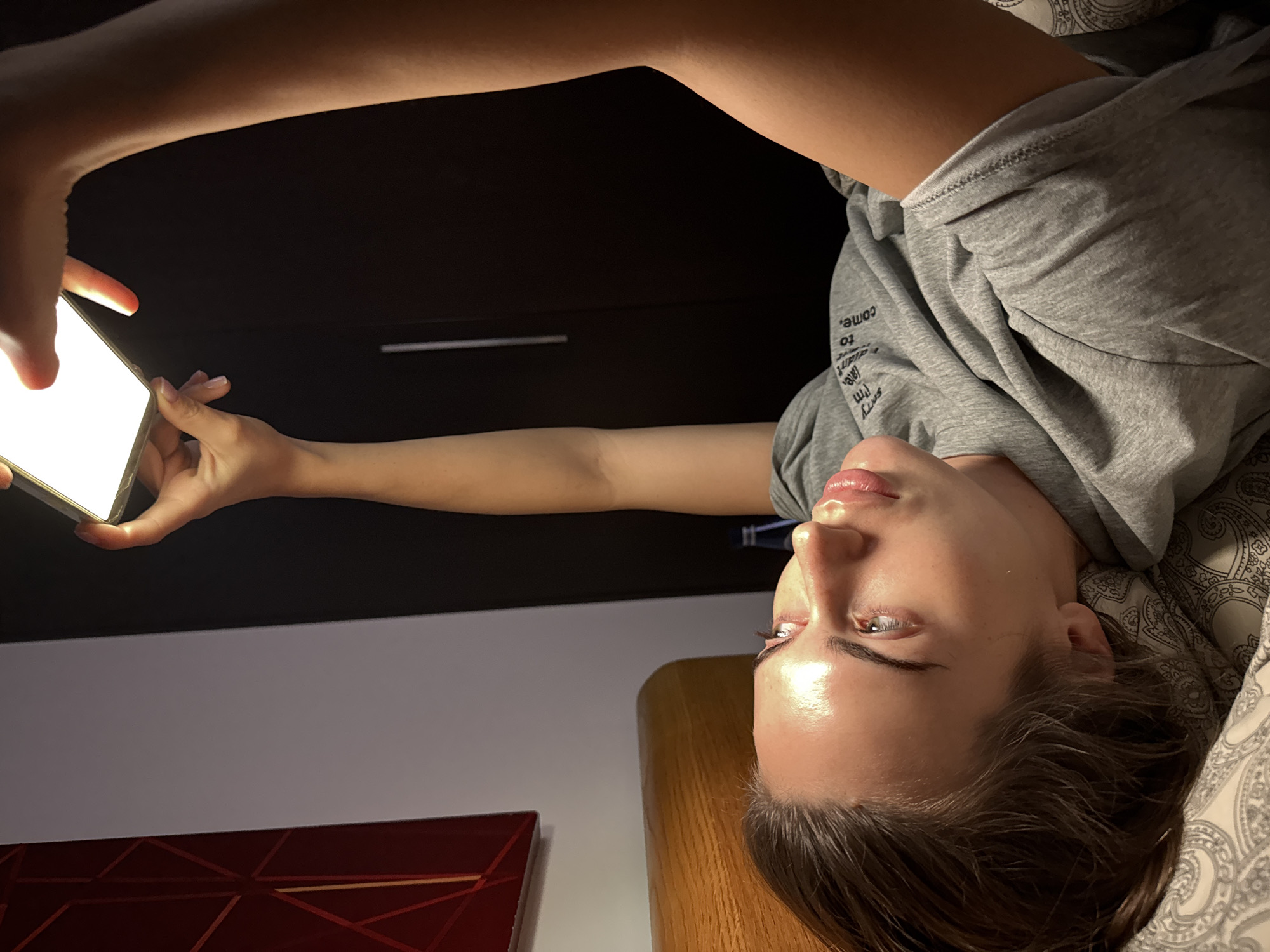 A person is lying down, looking at a brightly lit mobile device held in their hands. The person has fair skin, brown hair, and is wearing a gray hooded shirt. They are lying on a patterned pillow. Behind them are dark wood cabinets and a white wall above a reddish-brown object. There is no information to suggest a location in Warsaw, Poland.