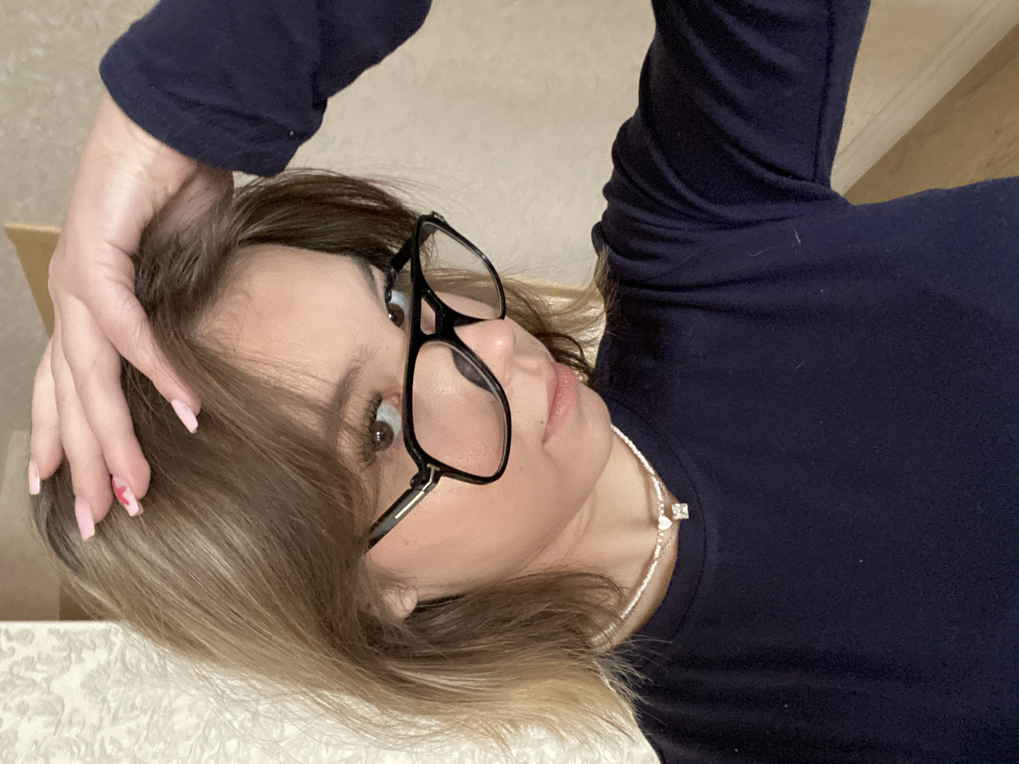 This image depicts a young female indoors, in the city of Chaikovsky, Russia. The subject has light brown hair with blonde highlights and wears black-framed glasses with gold accents. Her dark brown eyes are visible through the lenses. She is dressed in a dark navy blue long-sleeved shirt. Around her neck, she wears a layered necklace featuring a small white heart charm and a square, clear gemstone pendant. Her left hand is raised, with fingers splayed and resting on the top of her head, slightly parting her hair. Her fingernails are manicured with light pink polish, and a small red heart design is visible on one nail. The background shows an interior wall; to the left, a light-colored wallpaper with a subtle patterned design is visible, while to the right, a plain, lighter-toned wall with a partially visible wooden trim element is present. The subject is looking directly towards the camera.