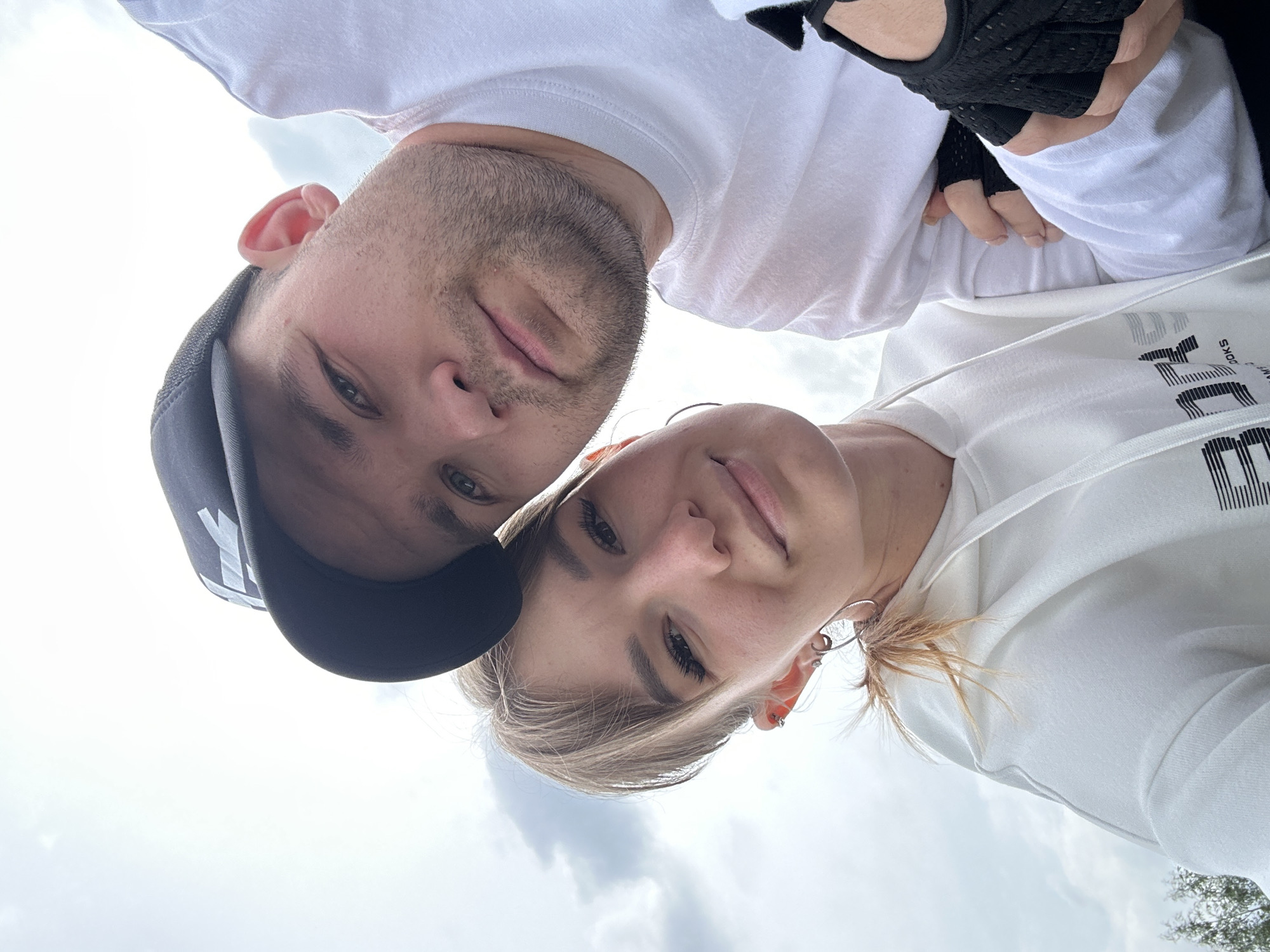 A close-up, low-angle shot captures two individuals, a male and a female, looking directly down at the camera. They appear to be taking a selfie outdoors in Chaikovsky, Russia, against a bright, partly cloudy sky.
The male subject is on the left, wearing a white t-shirt and a dark baseball cap with some indistinct white lettering on the front. He has short facial hair, and his right hand, partially visible, wears a black fingerless glove. He has a neutral to slight smile.
The female subject is positioned to the right of the male, wearing a white hoodie with the word "SPORT" clearly visible in dark lettering across the chest. She has light-colored hair, styled with some strands visible, and multiple earrings, including a large silver hoop. Her eyes are open, and she also displays a gentle, relaxed expression.
The background is dominated by a bright, overcast sky with visible clouds, indicating a daytime setting. A small sliver of green foliage, likely from a tree, is visible in the bottom right corner, confirming the outdoor environment. Both individuals are in close proximity, suggesting a shared moment or connection. They appear relaxed and content.