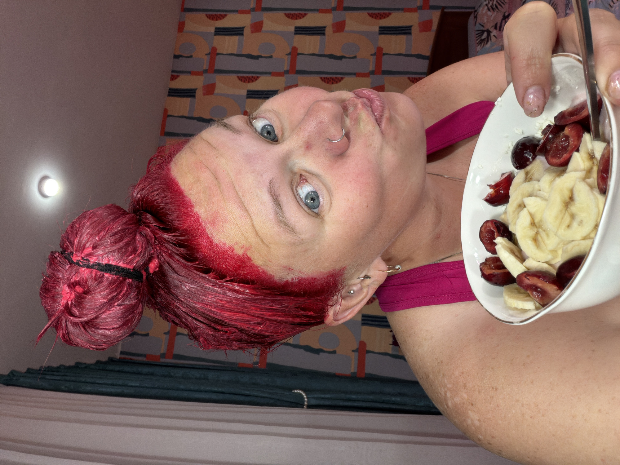 A woman with bright red hair, freshly dyed and showing red stains on her forehead and scalp, is captured in a candid, upward-facing shot. Her hair is styled in a bun, and she has piercing blue eyes, a nose ring, and multiple ear piercings, including a distinctive safety pin-shaped earring. She is wearing a fuchsia-colored top.
The woman is holding a white bowl filled with sliced bananas and dark red fruit, possibly cherries or plums, with a spoon visible within the fruit. Her gaze is directed towards the viewer, and her mouth is slightly open, conveying a casual or direct expression.
The background reveals an indoor setting with a patterned wall or curtain featuring geometric shapes in muted tones of grey, orange, and red. Below this, a dark teal fabric, possibly part of bedding, is visible. A circular light fixture shines brightly on a plain wall section to the upper left. The artificial light suggests an indoor, possibly evening environment. Her fingernails are painted with a glittery polish.