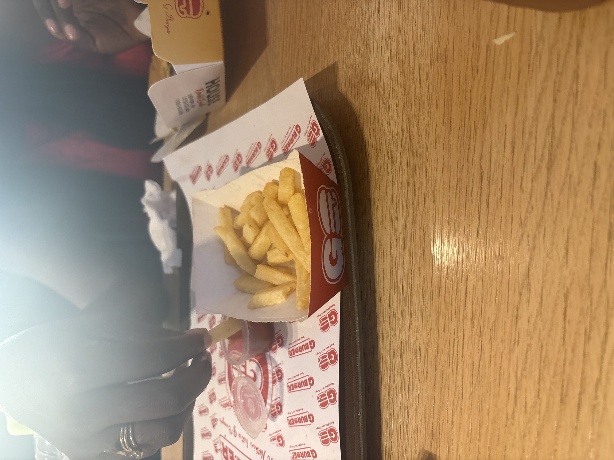 A tray of french fries and ketchup sits on a wooden table at G Burger, a fast-food restaurant in Abuja, Nigeria. A hand dips a fry into the ketchup. Part of a burger box is also visible.