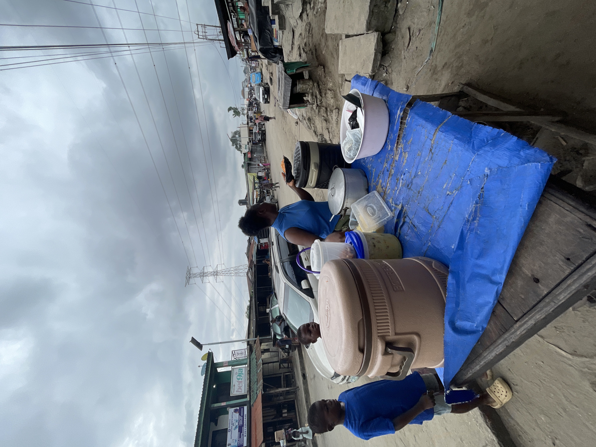 The image depicts a street scene in Ado, Nigeria, under an overcast sky. A woman, dressed in a blue sleeveless top, stands behind a table covered with a blue tarp, serving food. She is holding a black bowl containing what appears to be food, and a serving utensil. On the table, several food containers are visible, including a large beige insulated container, a large black insulated container, a metal pot, and a white plastic basin containing items. Next to these are smaller containers, one holding a yellow-green substance.
Two young boys are present. One, wearing a blue t-shirt and shorts, stands to the left of the table, looking towards the woman. The other boy, also wearing a blue t-shirt, is partially visible behind the beige insulated container.
The background shows a street with buildings lining it, some with signage indicating businesses. Vehicles, including a white SUV and a blue bus, are on the road. Overhead, power lines and a transmission tower are prominent. The ground is dirt and debris, with some concrete structures visible. The overall environment suggests an outdoor market or street-side vendor setup in an urban area.