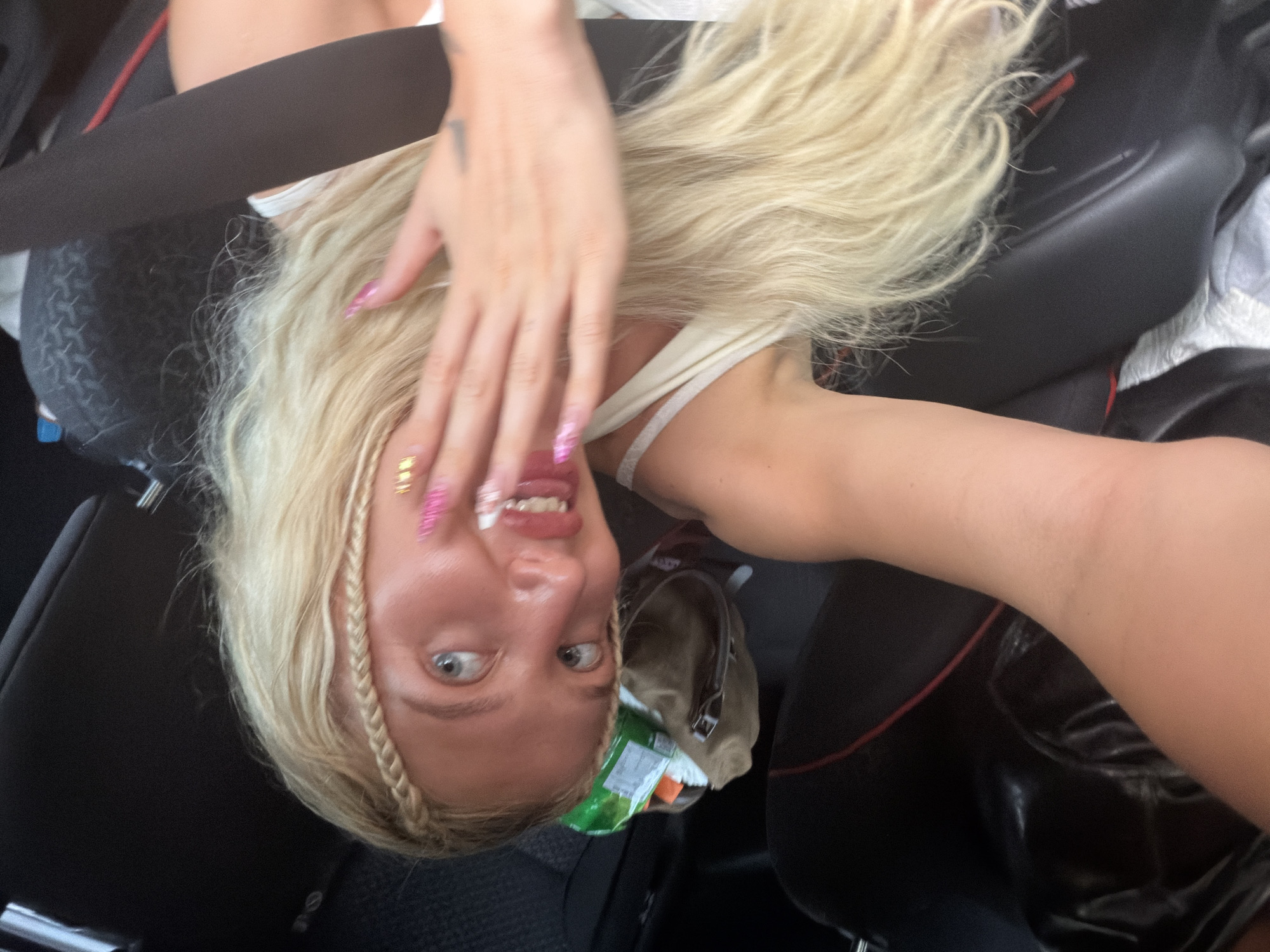 A young woman with blonde braided hair and blue eyes is sitting in the passenger seat of a car, holding her right arm up to her face. Her nails are long and painted with pink and gold glitter. She is wearing a white tank top. Her left arm is visible as well, resting on her lap. A duffel bag filled with snacks and other items is visible in the backseat behind her. The car appears to be parked. The scene is indoors, likely in a residential setting.