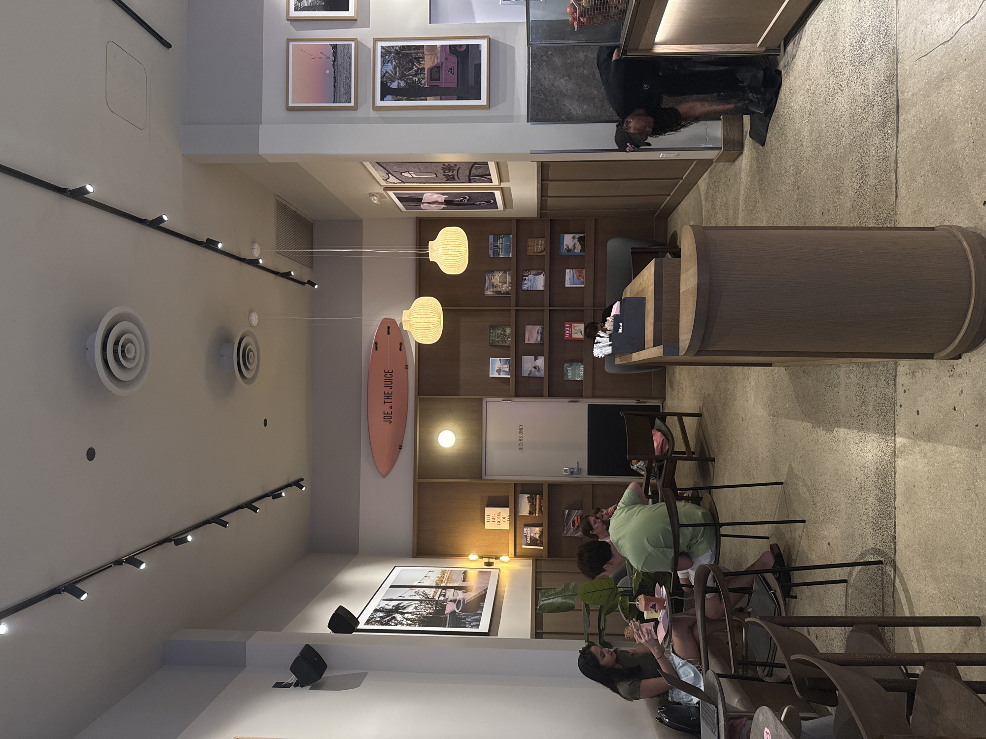 The image shows the interior of a Joe & The Juice location. There are customers sitting at tables and a Joe & The Juice employee. The location of this store in the city of Miami, United States of America cannot be determined from the given image.