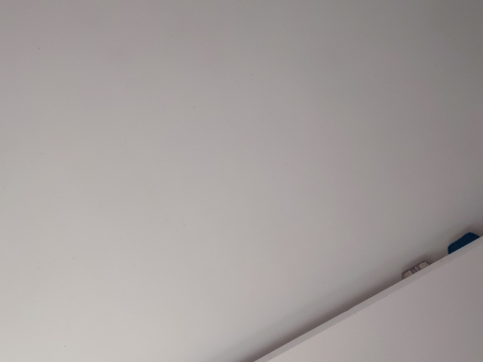 The image captures an indoor scene dominated by a large, plain, off-white or light gray surface, likely a ceiling or wall, which fills the majority of the frame. The surface appears smooth, with some minor, subtle speckling. A distinct diagonal line cuts across the bottom right portion of the image, marking the edge of another light-colored surface, possibly a wall, shelf, or cabinet. Along this edge, in the bottom right quadrant, several small, reflective, metallic objects are visible, suggesting hinges, brackets, or decorative elements. Further to the right, near the corner formed by the two surfaces, a small, dark blue object, potentially plastic or fabric, is partially discernible. The scene is evenly lit, indicating indoor lighting conditions. No people or specific activities are depicted. There is no visible text. The location is Santa Ana, Venezuela.
