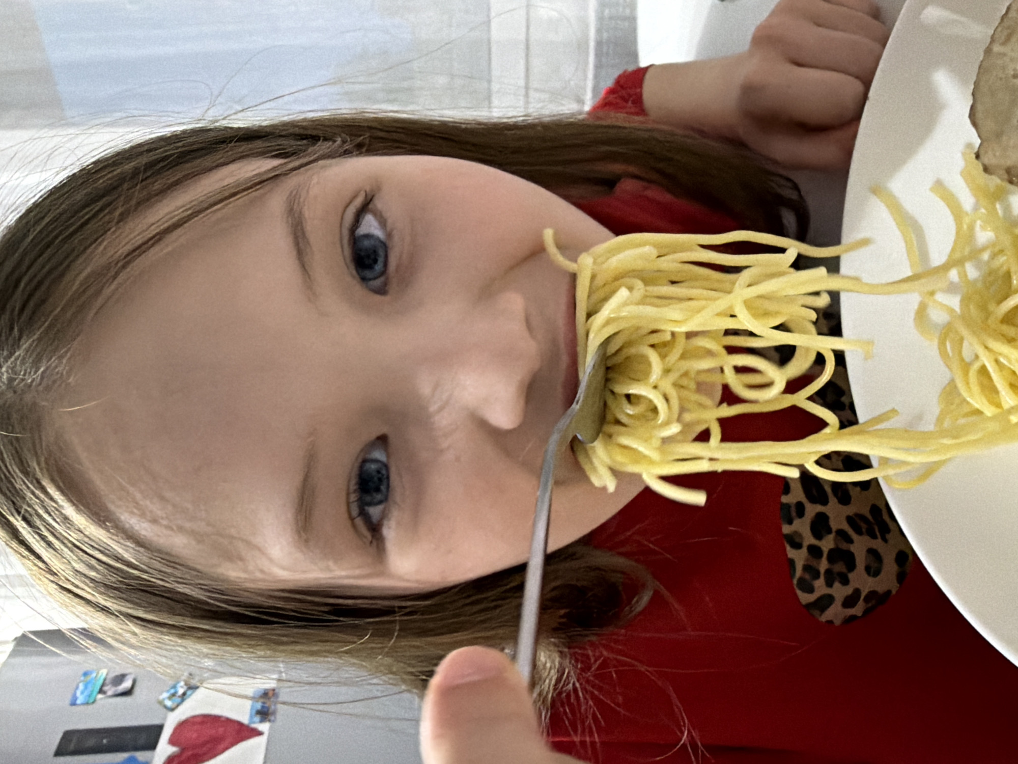 A girl is eating spaghetti with a fork. The girl has brown hair and blue eyes. She is wearing a red shirt with a black and brown leopard print collar. She is holding the fork in front of her mouth with spaghetti on it. There is a plate of spaghetti to the right of the girl. The background is blurry, but it appears to be a kitchen. Small pictures are visible on the wall in the background. The location is likely inside a home in Chaikovsky, Russia, based on the request context.