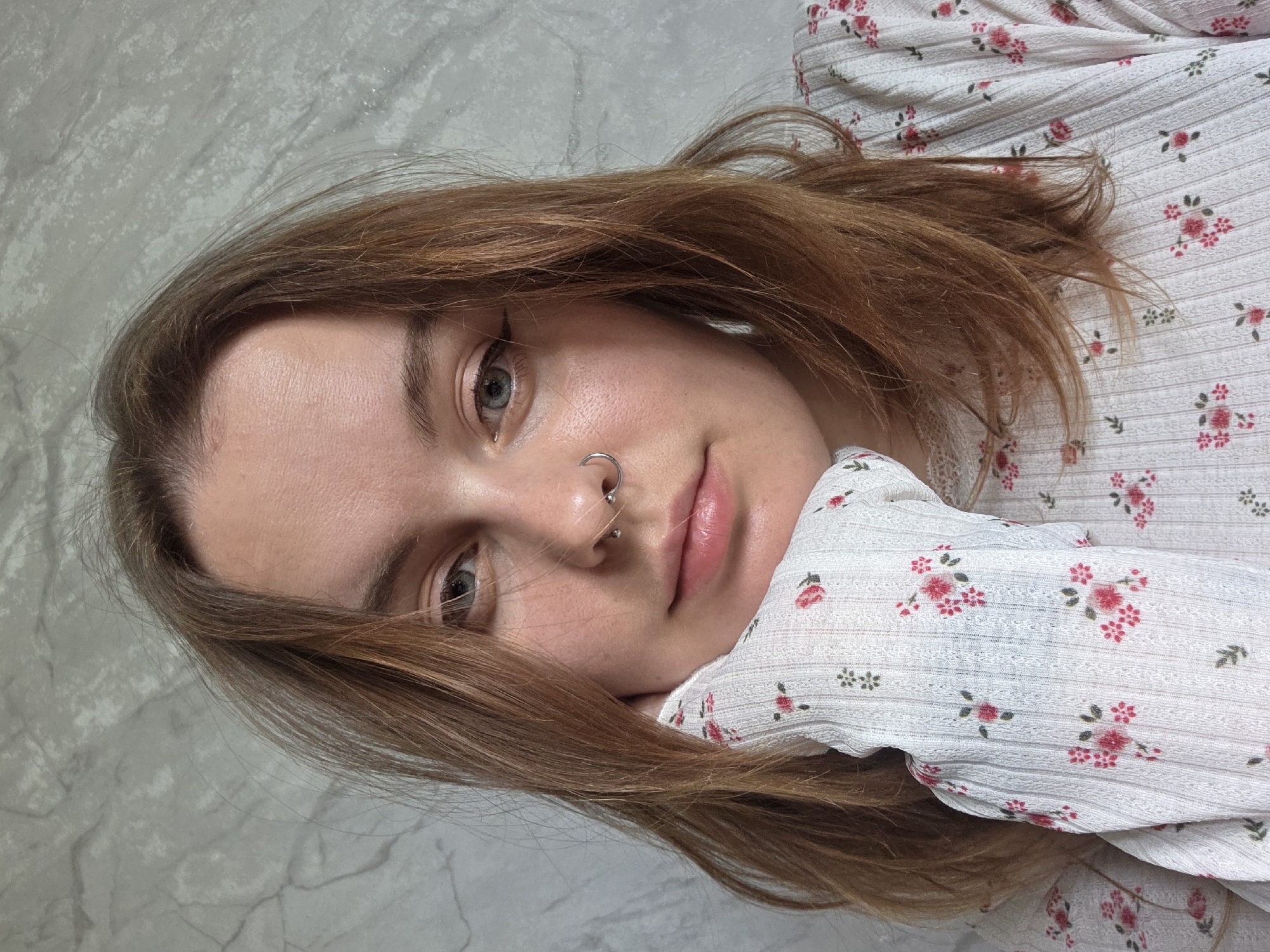 A close-up portrait of a young woman. Her face is partially turned towards the camera, revealing light brown hair with some reddish tones, and light skin. She has a nose piercing, and her eyes appear to be blue-gray in color. She is wearing a white floral-patterned top or dress. The background appears to be a light gray surface. The location is Unknown, Unknown.