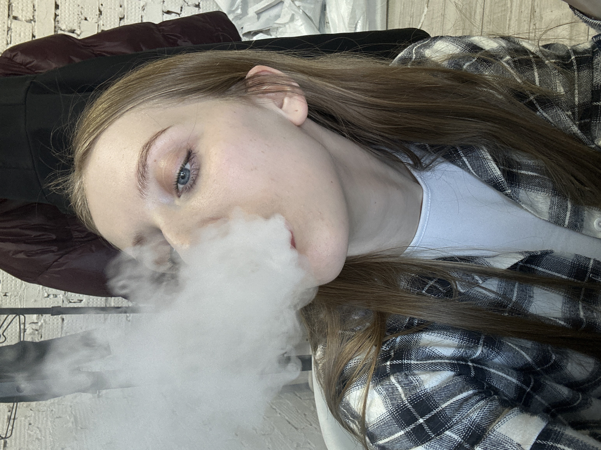 A female individual, estimated to be in her twenties, is depicted in an indoor setting. She has fair skin, blue eyes with visible eyeliner and mascara, and light brown hair. She is wearing a white t-shirt overlaid with a black and white plaid flannel shirt. A large, dense cloud of white vapor is actively being exhaled from her mouth, partially obscuring her left eye and nose. Her gaze is directed slightly upwards and to the left. The background consists of a white-painted brick wall. To the upper left and behind the vapor, a dark metal clothing rack holds various garments, including a dark jacket. A light-colored wooden or laminate floor is visible in the lower right portion of the frame. The specific location within Chaikovsky, Russia, is not identifiable from the visual information presented.