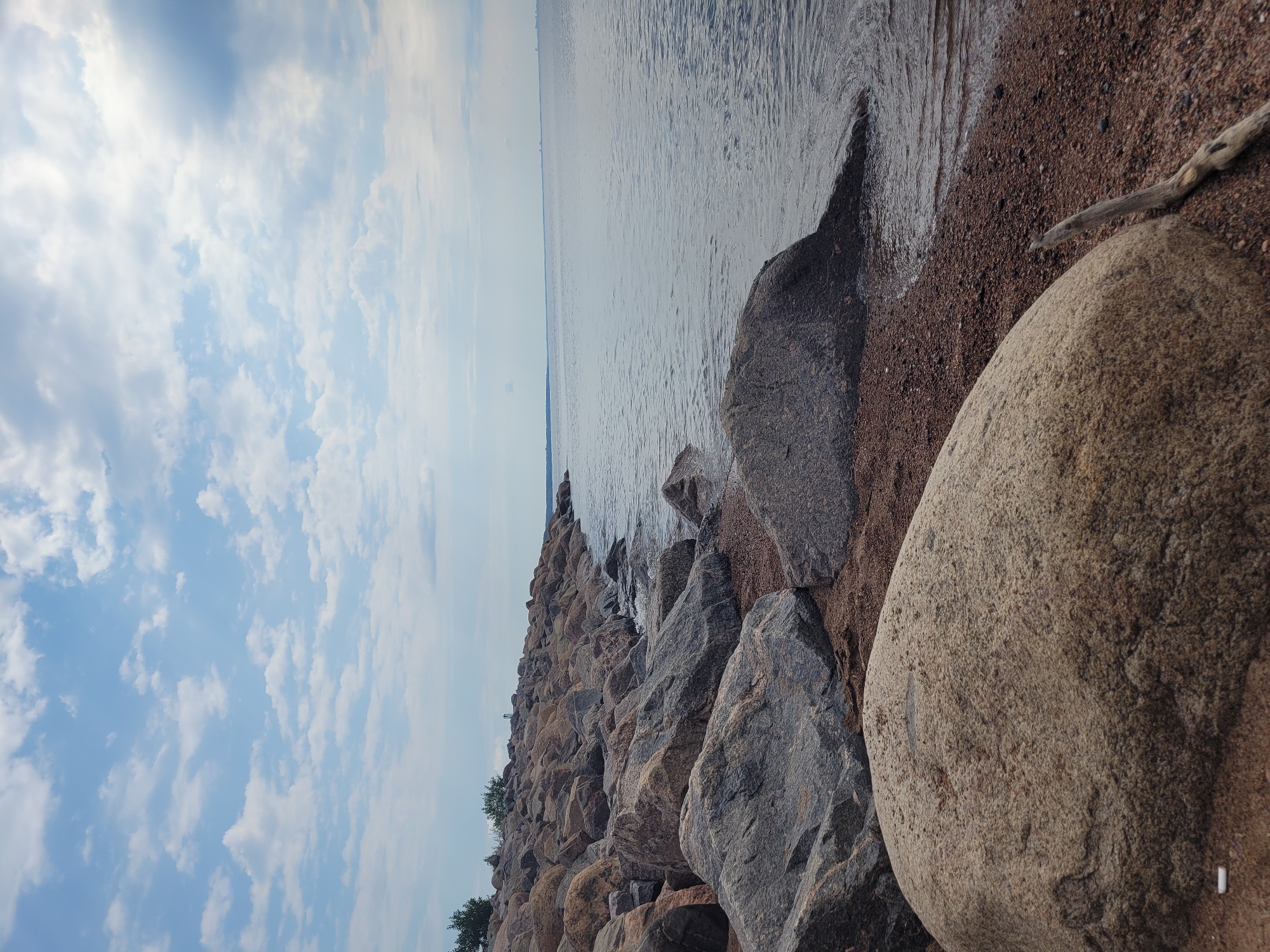 The image shows a rocky shoreline on the Gulf of Finland in Zelenogorsk, Russia. Large, weathered rocks of various sizes dominate the foreground and middle ground, some partially submerged in the calm, gray water. The shoreline is composed of small pebbles and sand. In the distance, the gulf stretches to the horizon under a partially cloudy blue sky with scattered white clouds. A faint line of trees is visible on the left side of the image along the upper edge of the rocky embankment. The scene appears to be a natural coastal landscape with no people or structures directly visible.