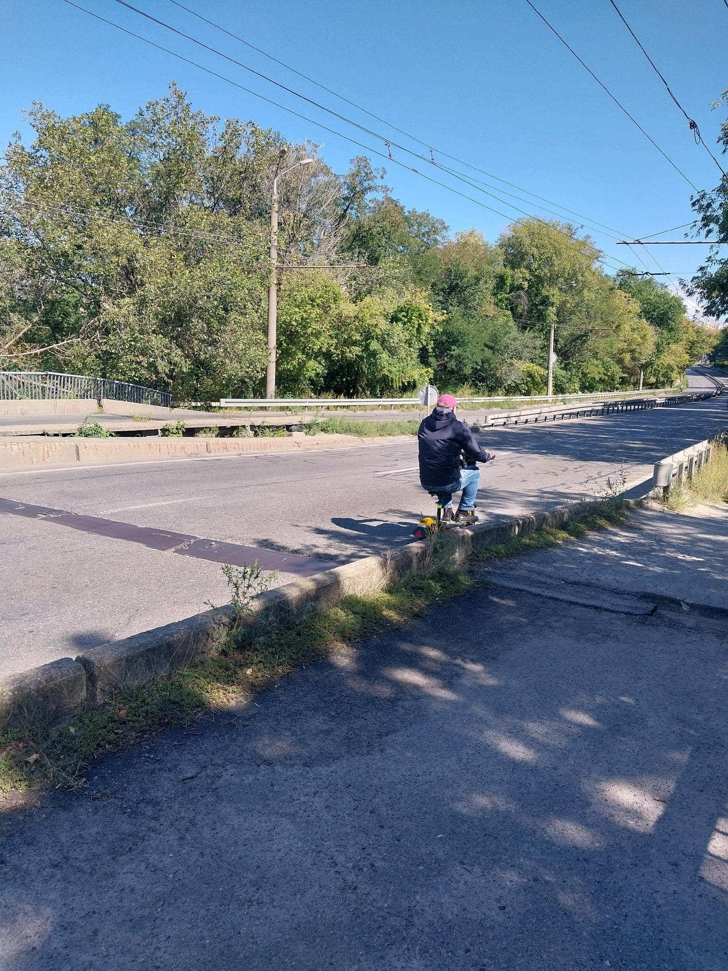 A person wearing a dark blue jacket, light blue jeans, and a pink cap is riding a yellow and black electric scooter on a paved road. The scooter has a black seat and a red light on the back. The person is facing away from the camera, heading down the road, which appears to be slightly inclined. The road has white lane markings and a concrete curb. To the left of the road, there is a pedestrian overpass with a metal railing. Trees with green foliage line the sides of the road, and overhead power lines are visible against a clear blue sky. The scene is likely located in Dnipro, Ukraine, given the presence of tram lines and typical urban infrastructure. The lighting suggests it is daytime, with shadows from trees cast on the road.