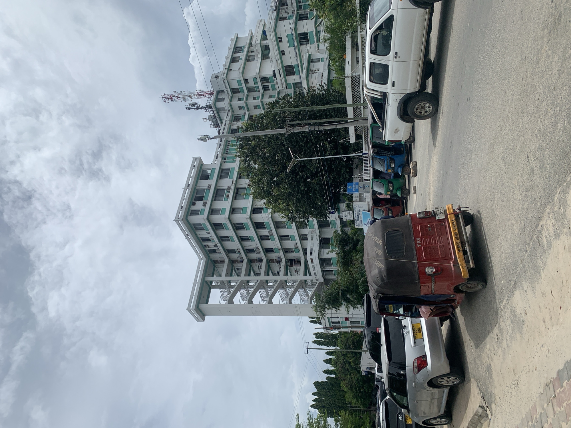 The image depicts a street scene in Dar es-Salaam, Tanzania. A multi-story, white building with green accents is visible in the background. The sky is overcast with grey clouds. In the foreground, there is a line of parked vehicles, including several cars and auto-rickshaws (tuk-tuks). A white pickup truck is also parked on the street. Utility poles with power lines are present, and trees are scattered throughout the scene.
