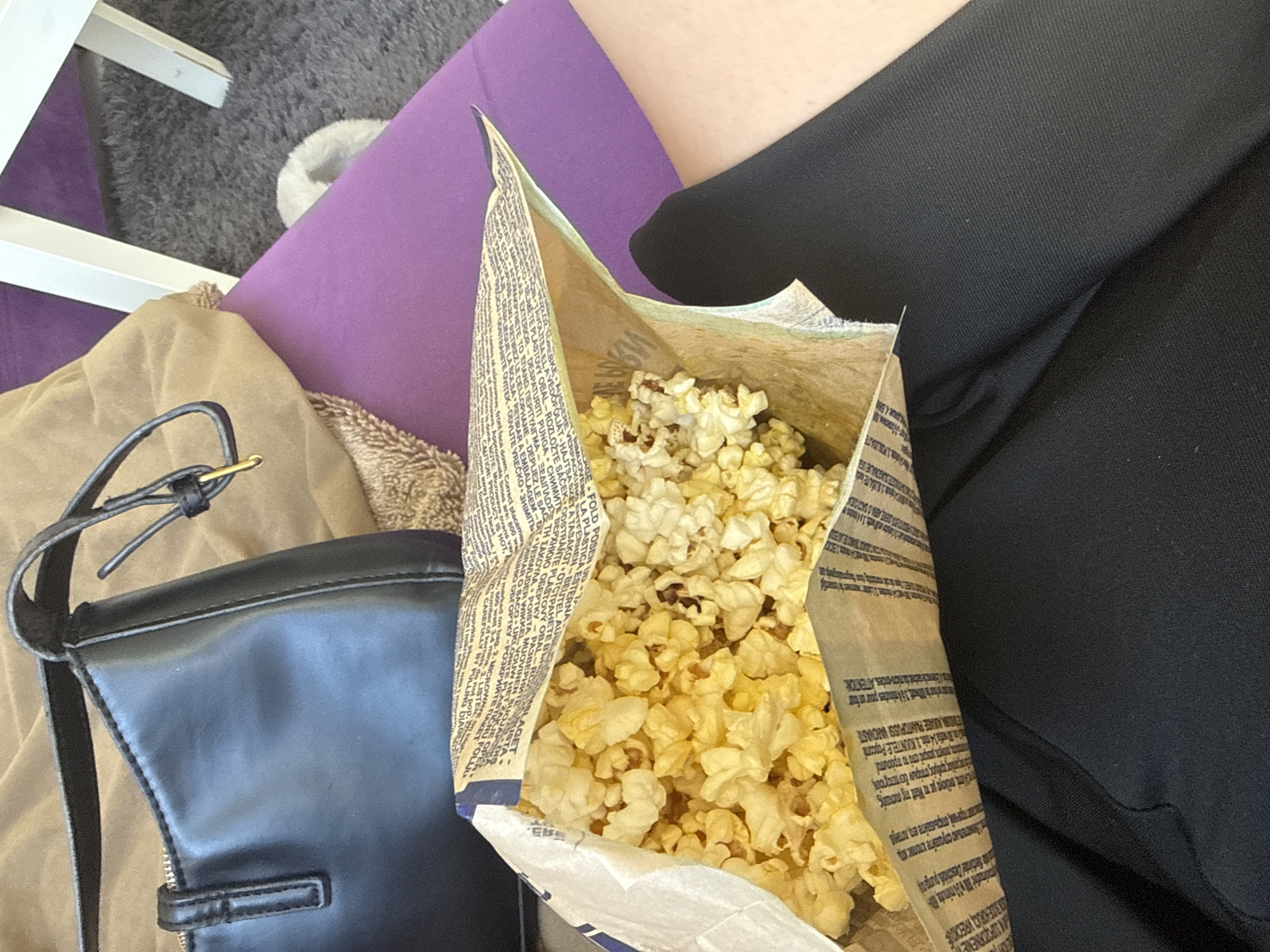 The image depicts a bag of popcorn sitting on what appears to be a purple cushion. Next to the popcorn is a black leather purse on a tan quilted surface. A leg covered in black material is also visible. A white chair leg is in the background. The text printed on the bag of popcorn is in several languages. The location is not identifiable as Sofia, Bulgaria, with the provided visual information.