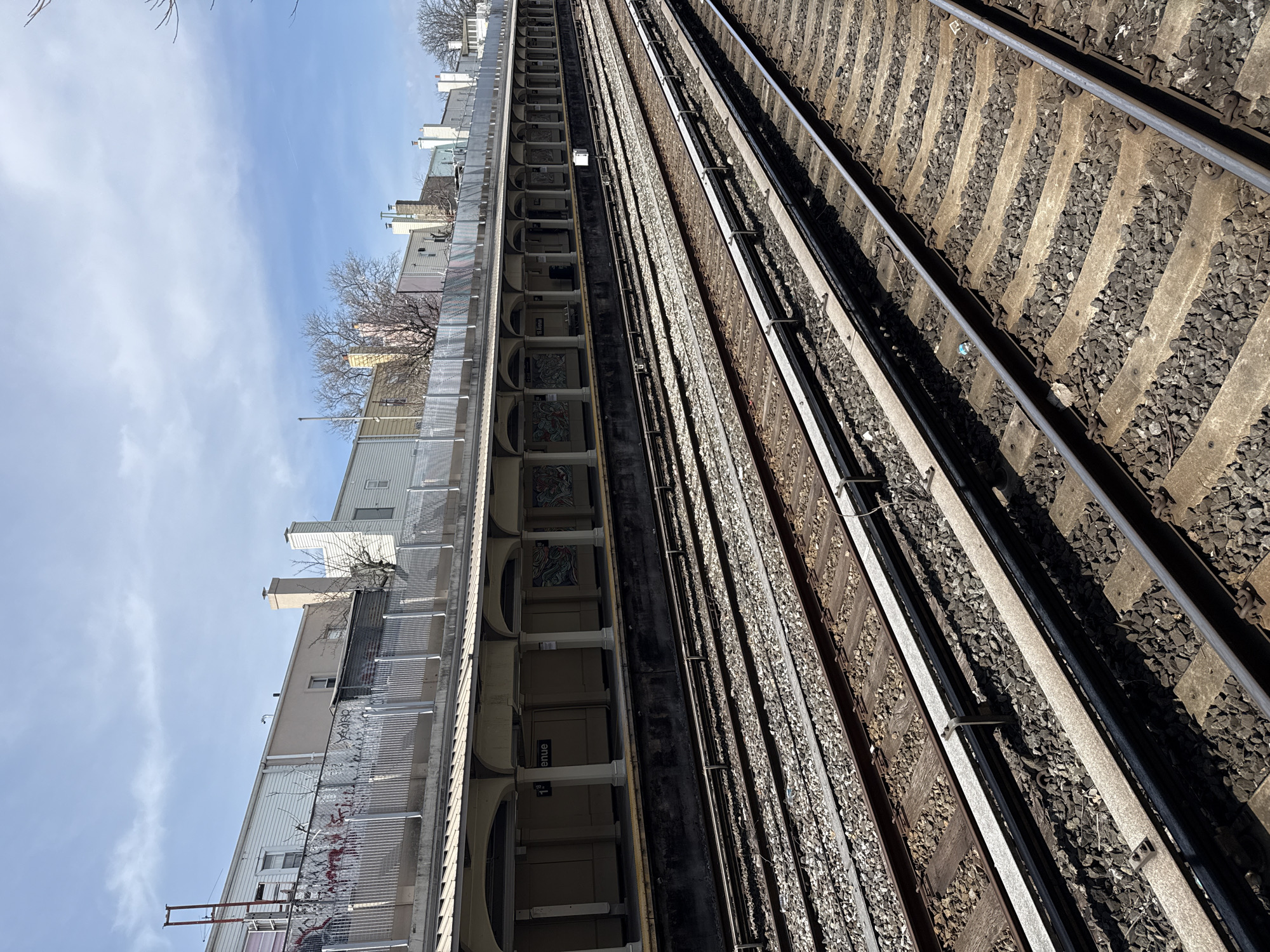 The image shows an elevated section of the New York City Subway. The focus is on the railway tracks running parallel to a station platform. The platform is characterized by architectural details like arched openings and tiled artwork. A sign indicating "18th Avenue" is visible. Adjacent to the station, multi-story buildings are seen, with some displaying graffiti. The sky is visible in the upper portion of the frame.
