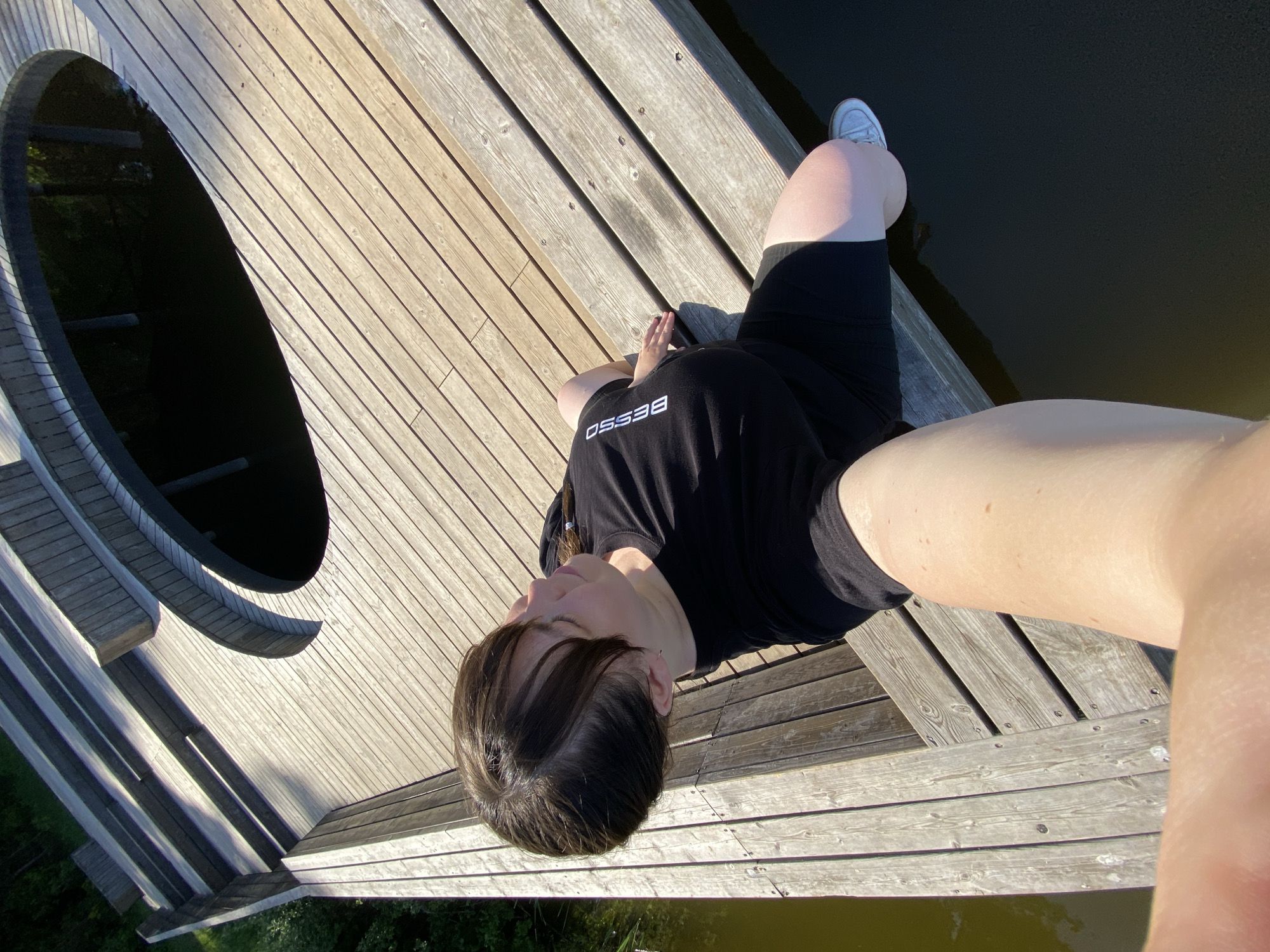 A young person with dark hair is sitting on a wooden structure next to a body of water. The structure appears to be a raised platform or bridge with a circular opening. The person is wearing a black t-shirt with white lettering, and black shorts. One white shoe is visible. The location is Votkinsk, Russia. The environment is sunny and outdoors.