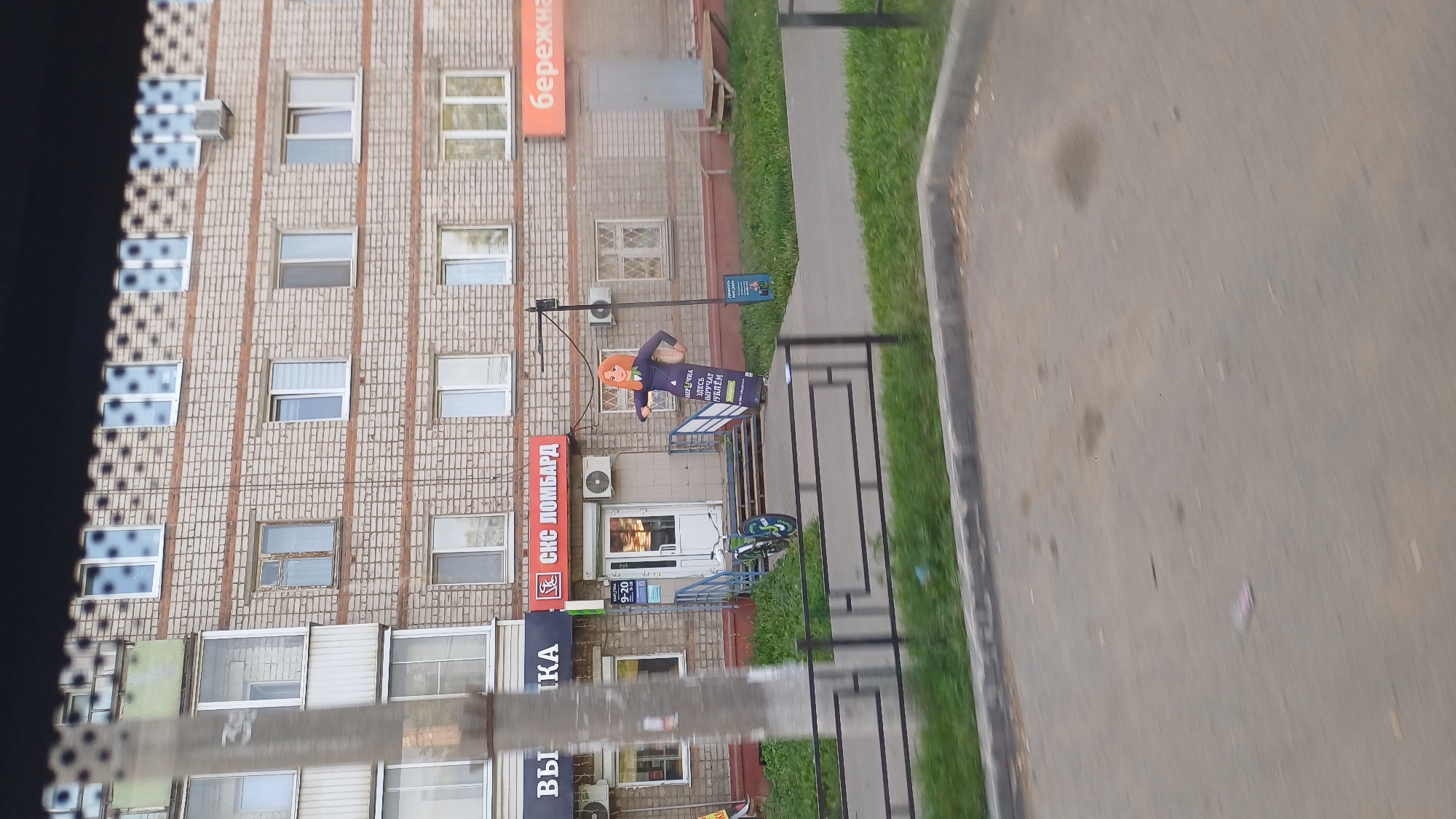 The image depicts a storefront in Izhevsk, Russia, characterized by a multi-story brick building with numerous windows. A prominent feature is a life-sized cardboard cutout of a woman in a business suit, holding a sign that advertises "МИР ЛИЧНОСТИ ЗДЕСЬ ПОЛУЧАТЬ РУБЛИ" (Personality World Here to Get Rubles). This cutout stands on the sidewalk in front of a pawn shop with the sign "СКС ЛОМБАРД" (SKS Lombard). Next to the pawn shop, another sign is visible, indicating operating hours from 9-20. A bicycle is parked on the stairs leading into the pawn shop. The scene includes a metal fence in the foreground, a grassy area, and a paved surface, likely a sidewalk or road. The overall impression is a commercial street view in a Russian city.
