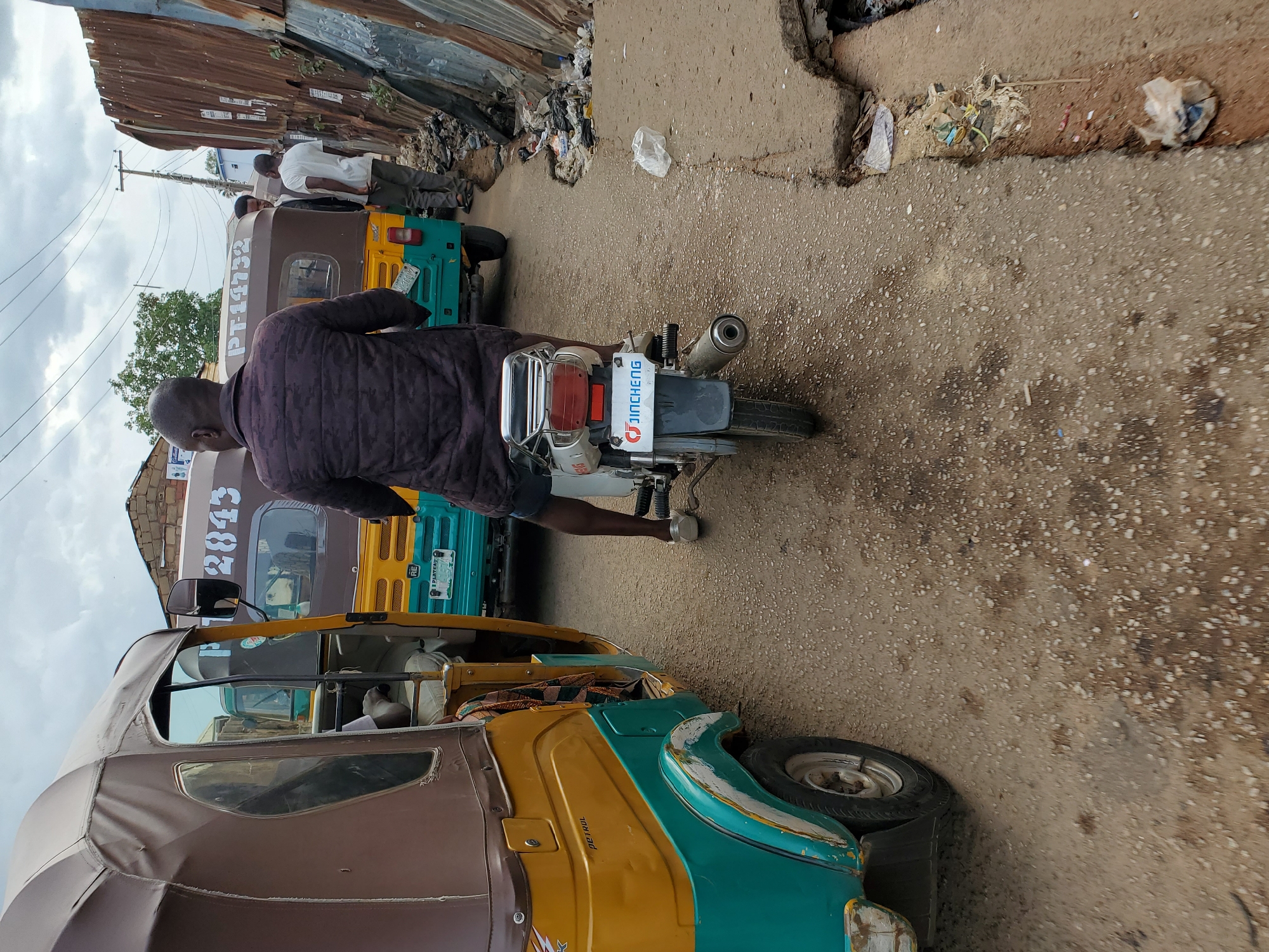 A street in Jos, Nigeria, is shown with three tricycles, one in the foreground, one in the middle ground, and one in the background. A man is standing next to a motorcycle in the center of the image. The street is narrow and cracked, with garbage visible on the edges. The buildings lining the street appear to be constructed of corrugated metal. The sky is overcast. The tricycle in the foreground is painted brown, yellow, and teal, while the tricycle in the middle ground is painted brown, yellow, and teal, with "P-14732" written on the back. The motorcycle in the center has a "JINCHENG" license plate. People are visible standing in the background.