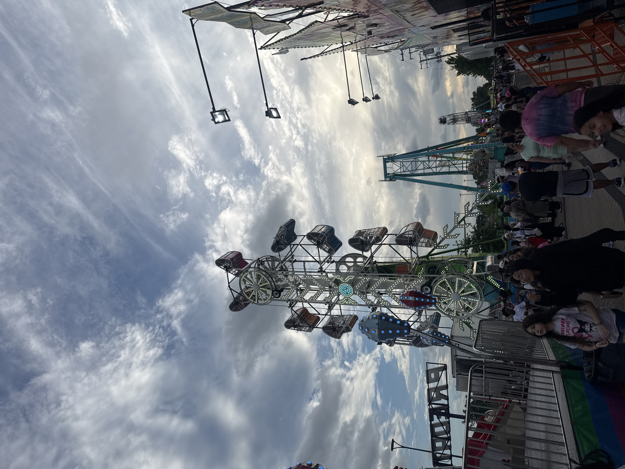 The image shows a busy street scene at a carnival in Niles, Illinois, USA. Several amusement rides, including a Ferris wheel and a pendulum ride, are visible. People are walking around and enjoying the attractions. The sky is cloudy, and the sun is setting, casting a warm glow over the scene. The overall atmosphere is festive and lively.