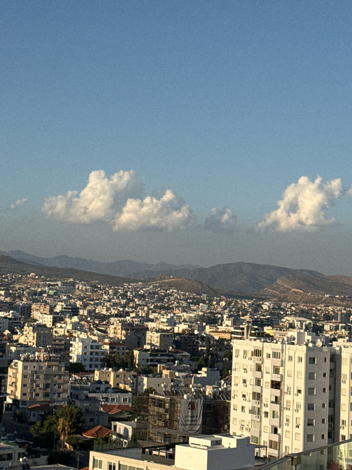 This is a high-angle, long shot of the cityscape of Limassol, Cyprus. The image captures a dense urban area with numerous buildings of varying heights, mostly appearing to be residential or commercial properties. The architecture is predominantly modern, characterized by concrete structures, balconies, and numerous windows. Many buildings have air conditioning units attached to their facades.
In the foreground, a modern, multi-story white building with a grid-like pattern of windows and balconies dominates the right side of the frame. Further to the left, other buildings of similar scale and design are visible. In the mid-ground, a vast expanse of smaller buildings creates a textured urban landscape. Red-tiled roofs are visible interspersed among the concrete structures.
The background features rolling hills and mountains, their shapes softened by distance and possibly atmospheric haze. The lower slopes of the hills show some vegetation, and some scattered buildings appear to be built on these slopes as well.
The sky above is a clear, pale blue, with a few scattered, fluffy white clouds. The lighting suggests it is either early morning or late afternoon, with a warm, diffused sunlight casting long shadows and illuminating the buildings. There are no visible people or vehicles in the image. The overall impression is one of a bustling, developed city nestled against a natural, mountainous backdrop.