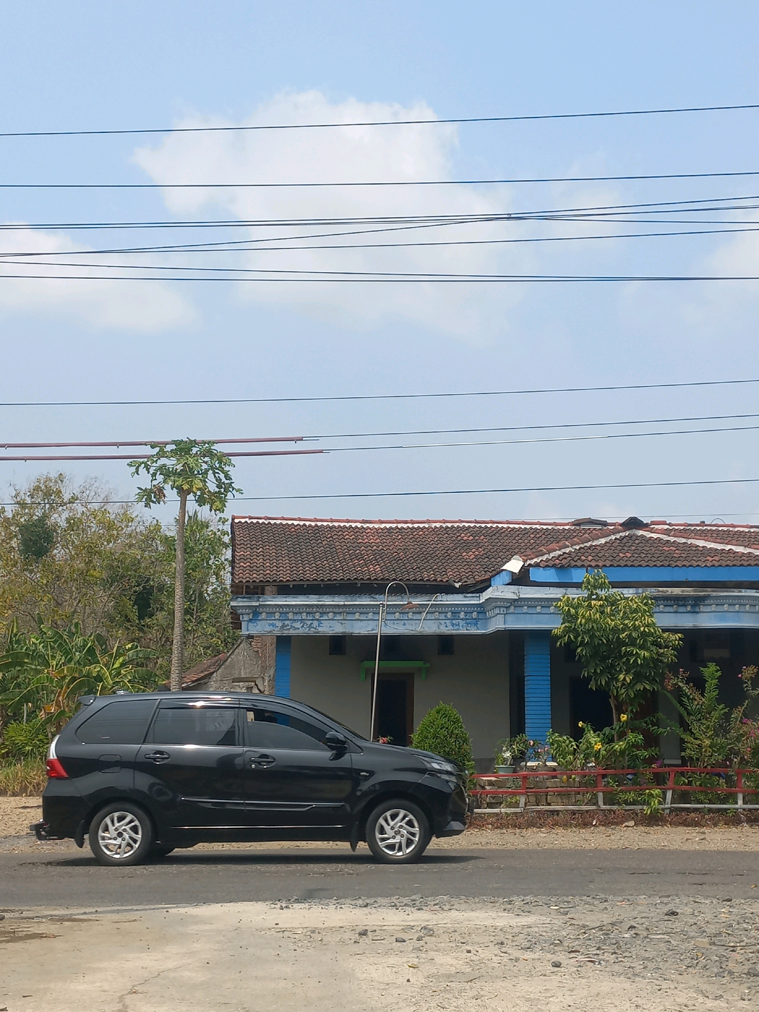 The image shows a black car driving on a road in front of a house. The house has a tiled roof and blue accents. There are trees and plants around the house. The sky is blue with some clouds. The scene appears to be in a rural or suburban area, consistent with the city of Baturetno, Indonesia. There are power lines running across the top of the image. The car is seen from the side, and it is the primary subject of the image. The road surface is a mix of asphalt and gravel.