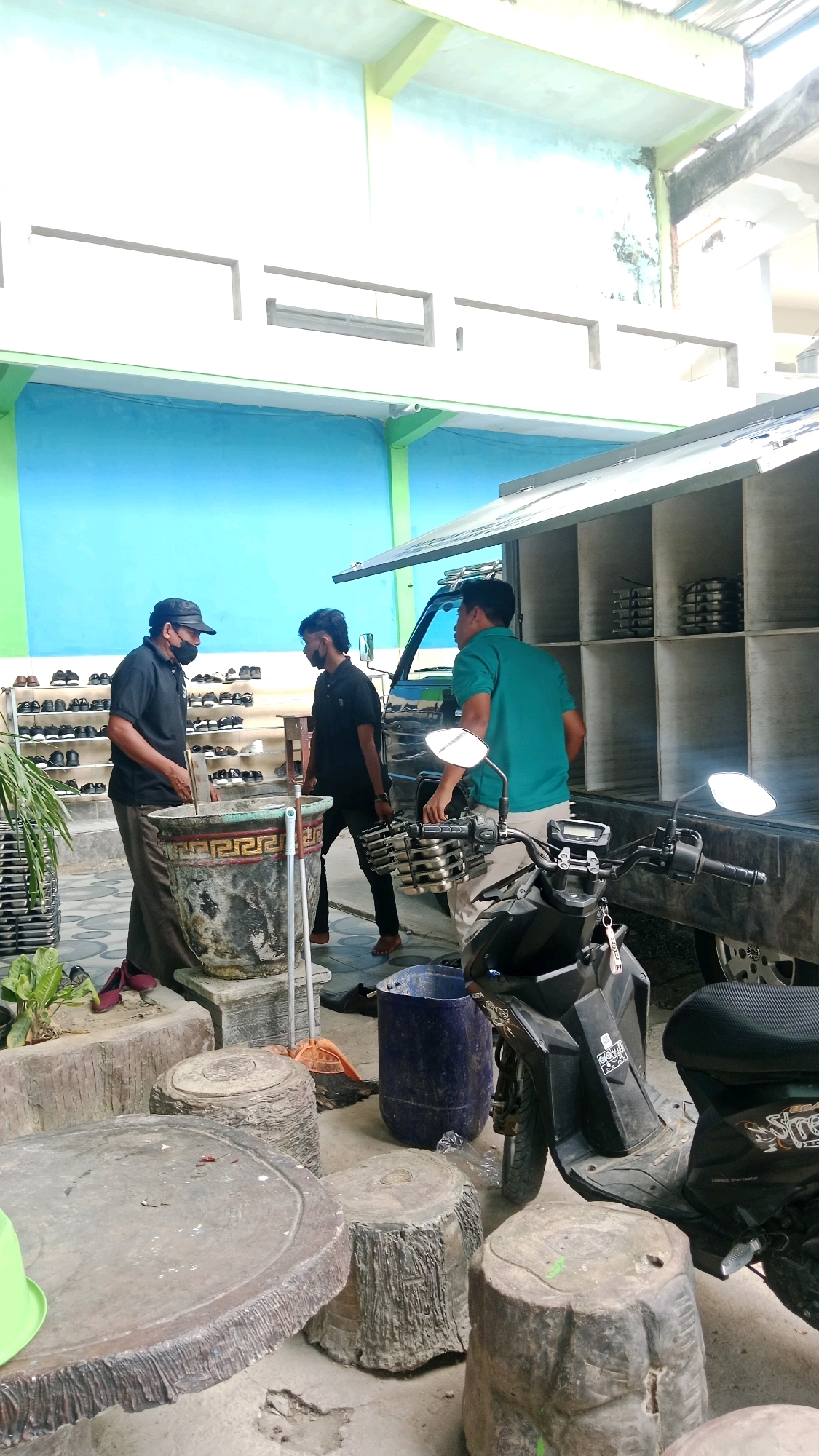 Three men are standing near a large, weathered concrete planter in what appears to be an outdoor area in Waru, Indonesia. One man, wearing a black cap and mask, stands facing the planter and gesturing with his left hand. To his right, a younger man in a black shirt also faces the planter, holding what appears to be a metal rack with circular objects. A third man, in a green shirt, stands to the right of the planter, facing away from the camera and towards the back of a black truck. The truck has an open rear section with shelves containing stacked metal items. A black scooter is parked to the right of the men, with its front wheel and handlebars visible. In the background, a wall painted blue and white can be seen, with a green trim along the bottom. Racks of shoes are visible to the left of the planter. The ground is concrete, with several large, rough-textured concrete seats or tables in the foreground. A blue plastic bin and an orange broom are also present on the ground. The scene suggests a communal or public space, possibly near a mosque or other religious building given the shoe racks.