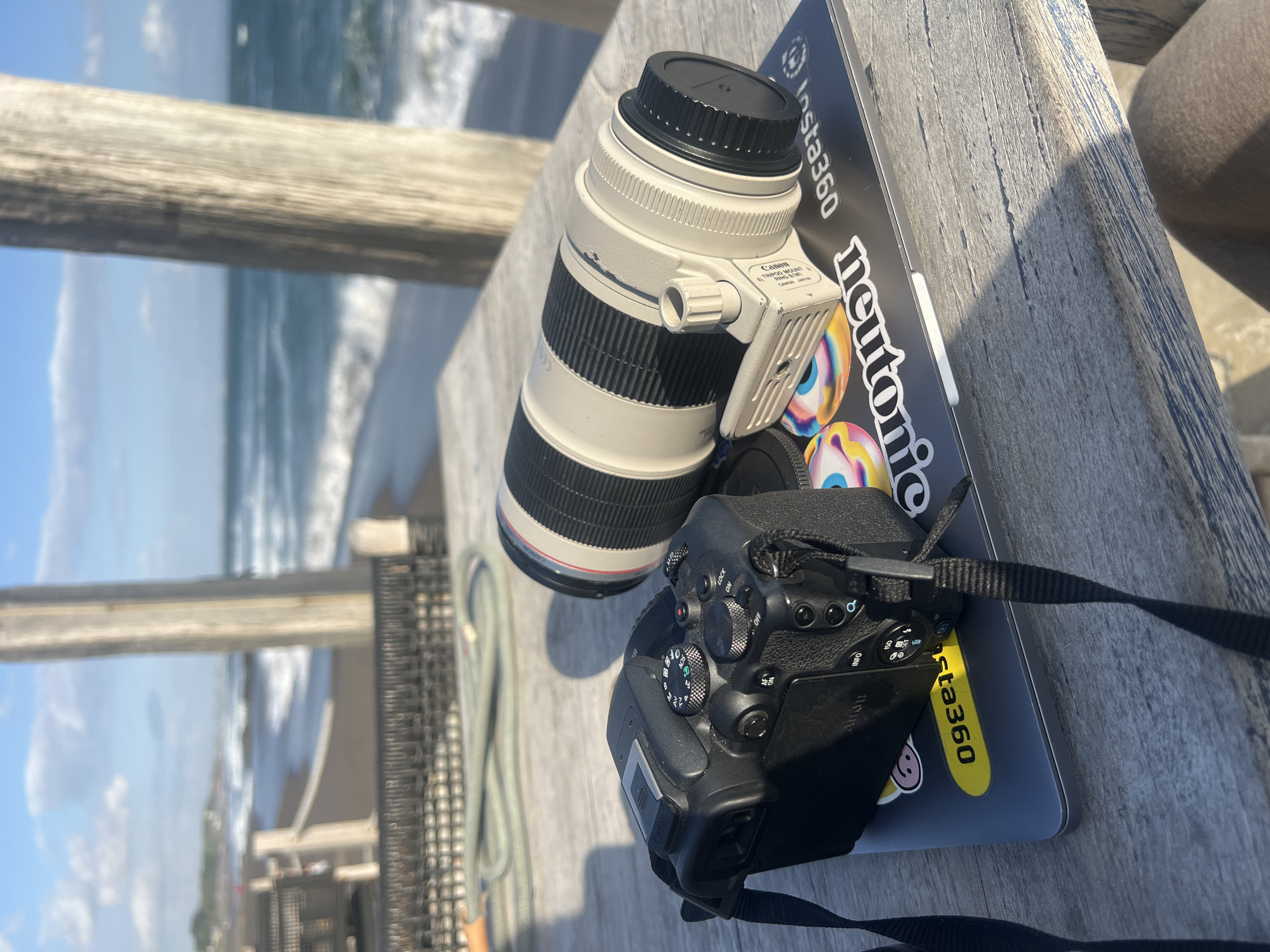 This is a photograph of camera equipment, specifically a Canon telephoto lens and a DSLR camera, resting on a wooden table outdoors. The camera and lens are placed on top of a laptop, which is adorned with stickers, including one that says "insta360" in white text on a black background, and another with colorful eye graphics and the text "newtonic" underneath. The DSLR camera has its strap attached and visible.
The scene is set outdoors, with a blurry background showing the ocean and waves under a bright sky, suggesting a beach or coastal location. The lighting indicates it is daytime, possibly late morning or afternoon, given the sunlight casting shadows. The weathered wooden surface of the table and a wooden railing in the foreground provide location cues. There is no clear indication of people or specific activities other than the setup of photographic equipment, implying the user might be preparing to take photos or has just finished. The overall mood seems relaxed and focused on the gear, perhaps anticipating a scenic shoot.