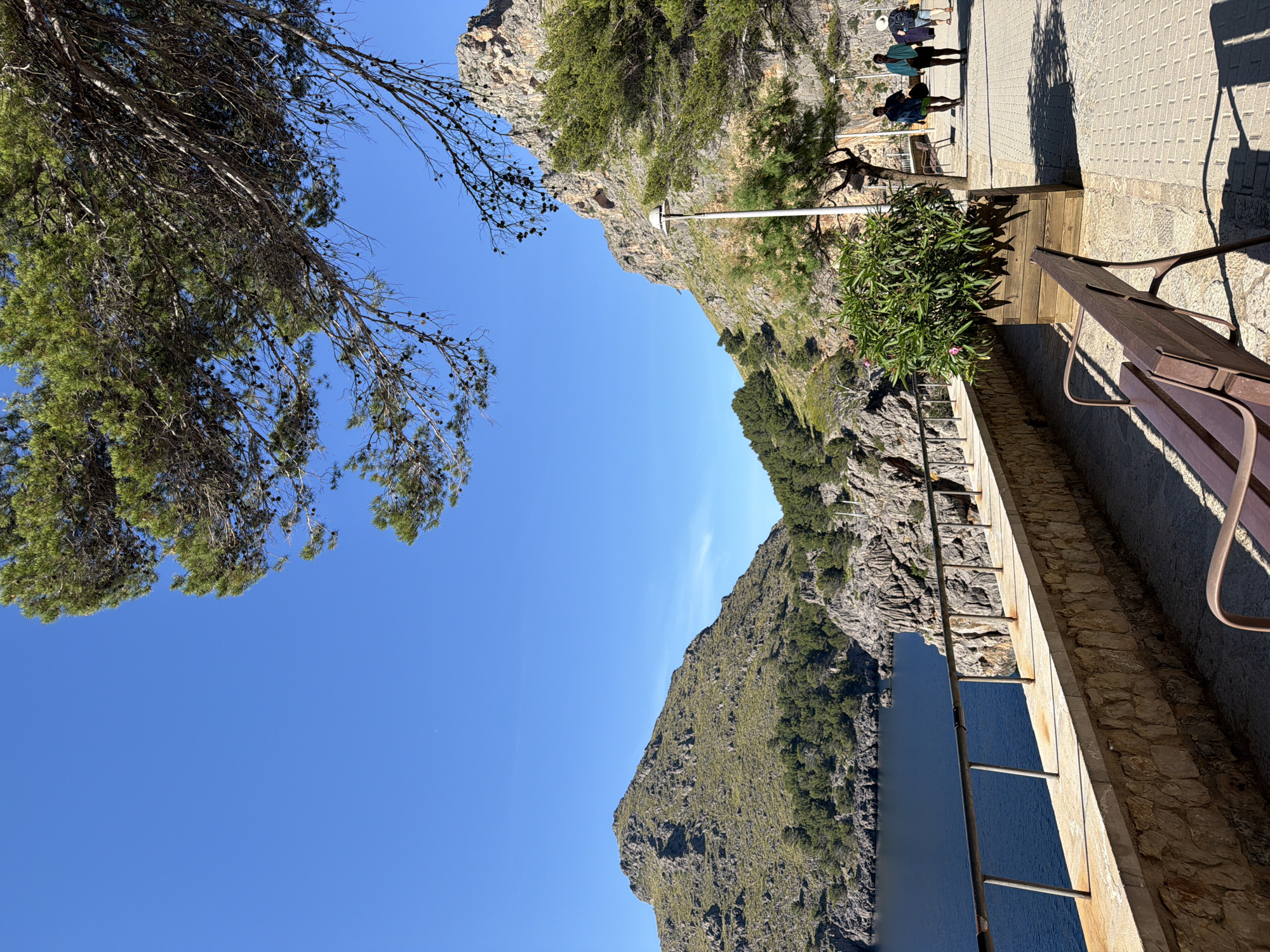 The image depicts a scenic overlook along the coast of Escorca, Spain. The foreground features a stone wall with a metal railing, along which a park bench sits. To the right, three people stand on a paved walkway, observing the view. Beyond the wall and railing, a steep, rocky cliff face descends to a body of water, likely the Mediterranean Sea. The cliff is partially covered in vegetation. In the upper left, tree branches frame the scene against a clear blue sky.
