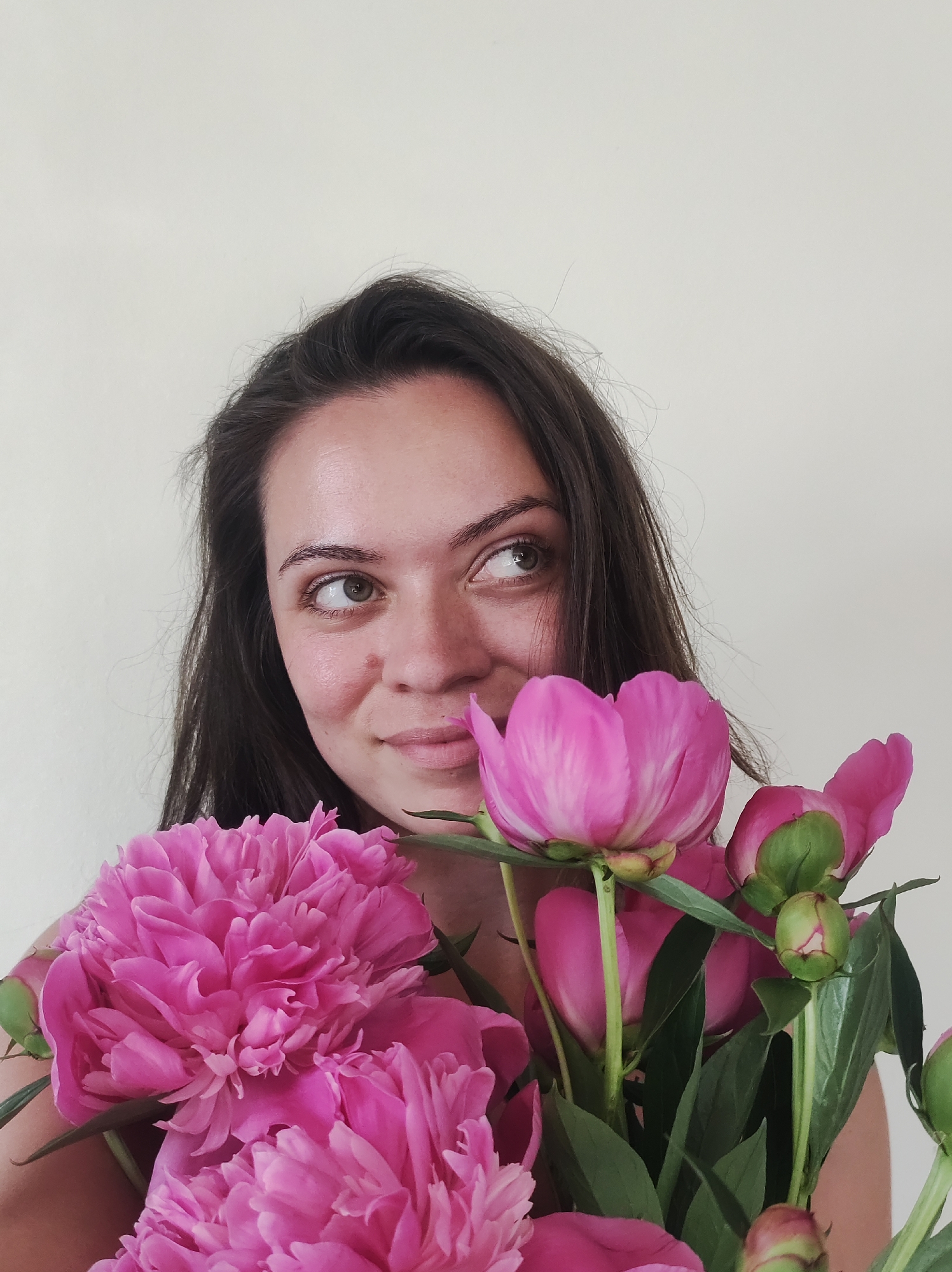 A medium shot, eye-level, portrait-style photograph shows a young woman with long, dark brown hair and light skin holding a bouquet of bright pink peonies. She is positioned in the lower left quadrant of the image, looking to the upper right with a slight smile. Her gaze is directed away from the camera, obscuring one eye with the flowers. The peonies are prominently displayed in the foreground, with several fully bloomed flowers and some buds. The background is a plain, light-colored wall.