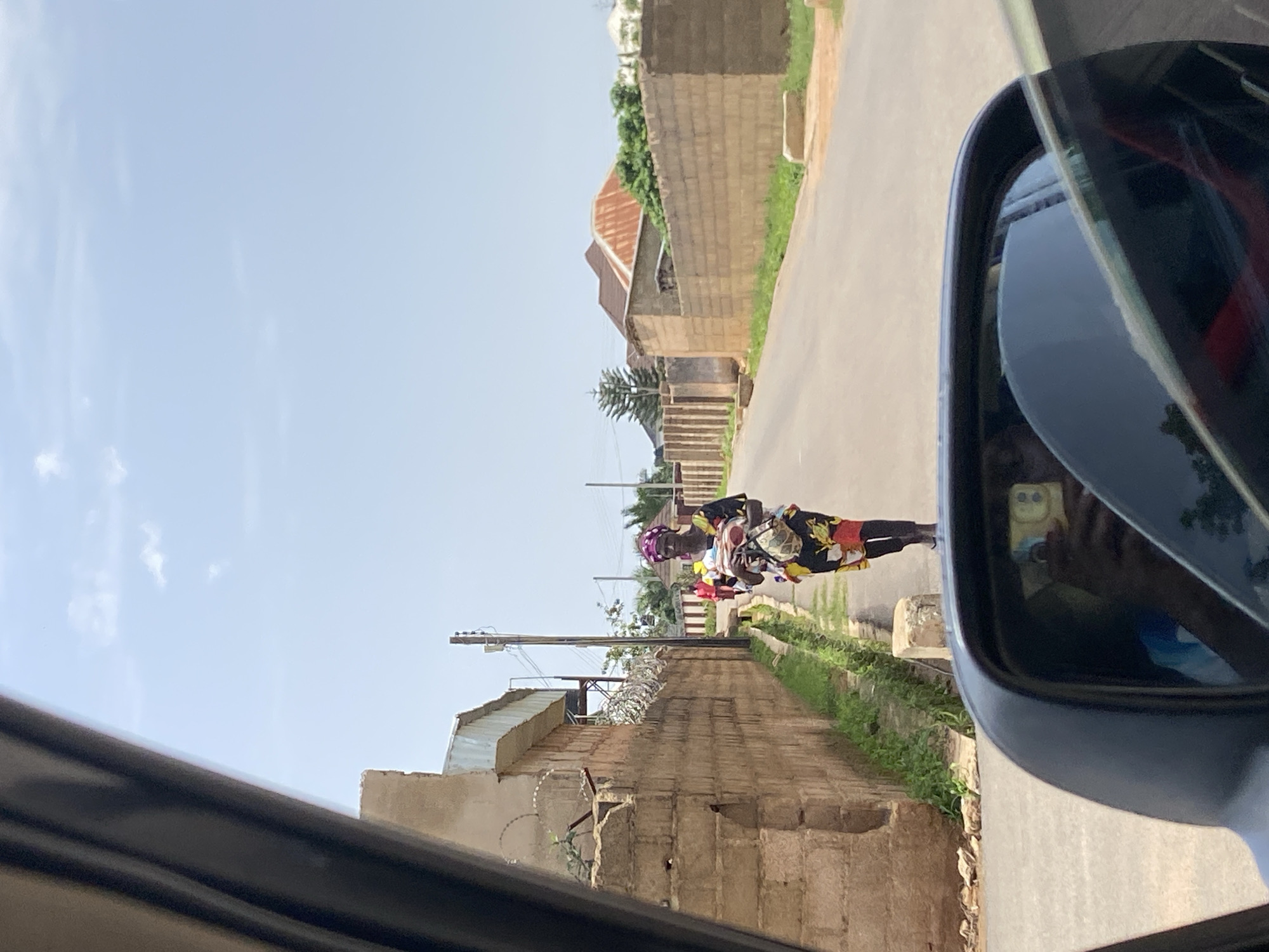 The image shows a street scene in Bar Jirgi Summa, Nigeria. In the foreground, the reflection of a person holding a smartphone is visible in a car's side mirror. The street is paved, with a grassy verge and a low wall on the left side, followed by a brick wall with barbed wire on top. On the right side of the street, there is another brick wall and various buildings with corrugated metal and tiled roofs in the background. A woman in a colorful dress and headscarf is walking along the street, carrying items wrapped in fabric. Another person in a red shirt is walking further down the street. The sky is clear and blue with a few scattered clouds.