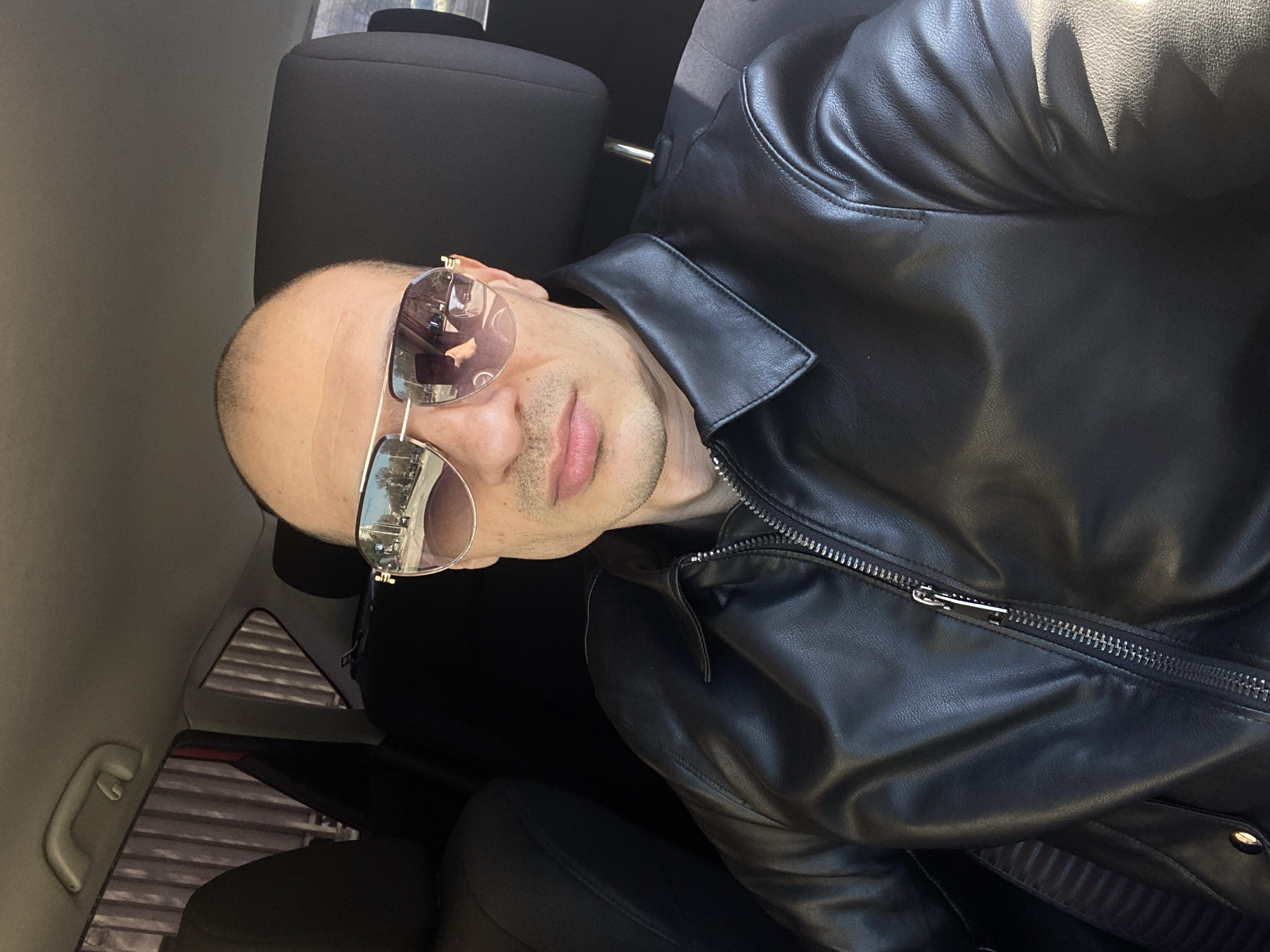 A bald man wearing sunglasses and a black leather jacket is seated in a car. The background includes parts of the car's interior, such as the seat, headrest, and window. The reflection in the sunglasses shows an urban environment, possibly a street scene in the city of Pleven, Bulgaria, based on the description.