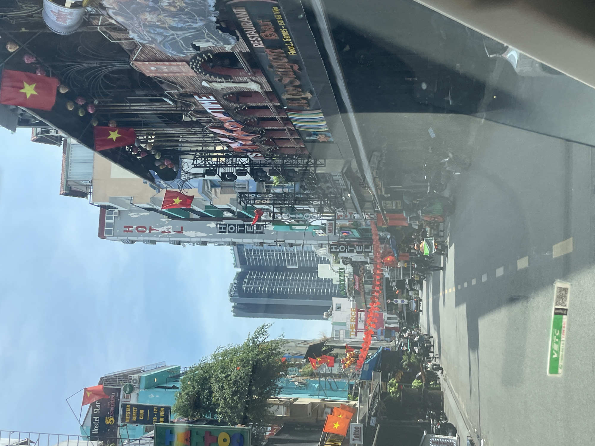 In Thủ Đức City, Vietnam, a street scene unfolds under a bright sky. The camera captures a perspective from a vehicle, likely looking forward through a windshield. The street is lined with buildings of varying heights and architectural styles, many adorned with Vietnamese flags. Businesses such as "Hostel Star," "Vietstar Buffet Club," and "Hotel" are visible on signs. A prominent building features a large sign reading "Miss Saigon Restaurant Bar," indicating its establishment on Floor 1, Games, 15B Bui Vien. Red festive decorations hang across the street in some sections. Several motorcycles and scooters are parked along the roadside and in motion on the street. A few individuals are present, walking or riding. The environment appears to be an urban setting with commercial establishments and residential buildings.