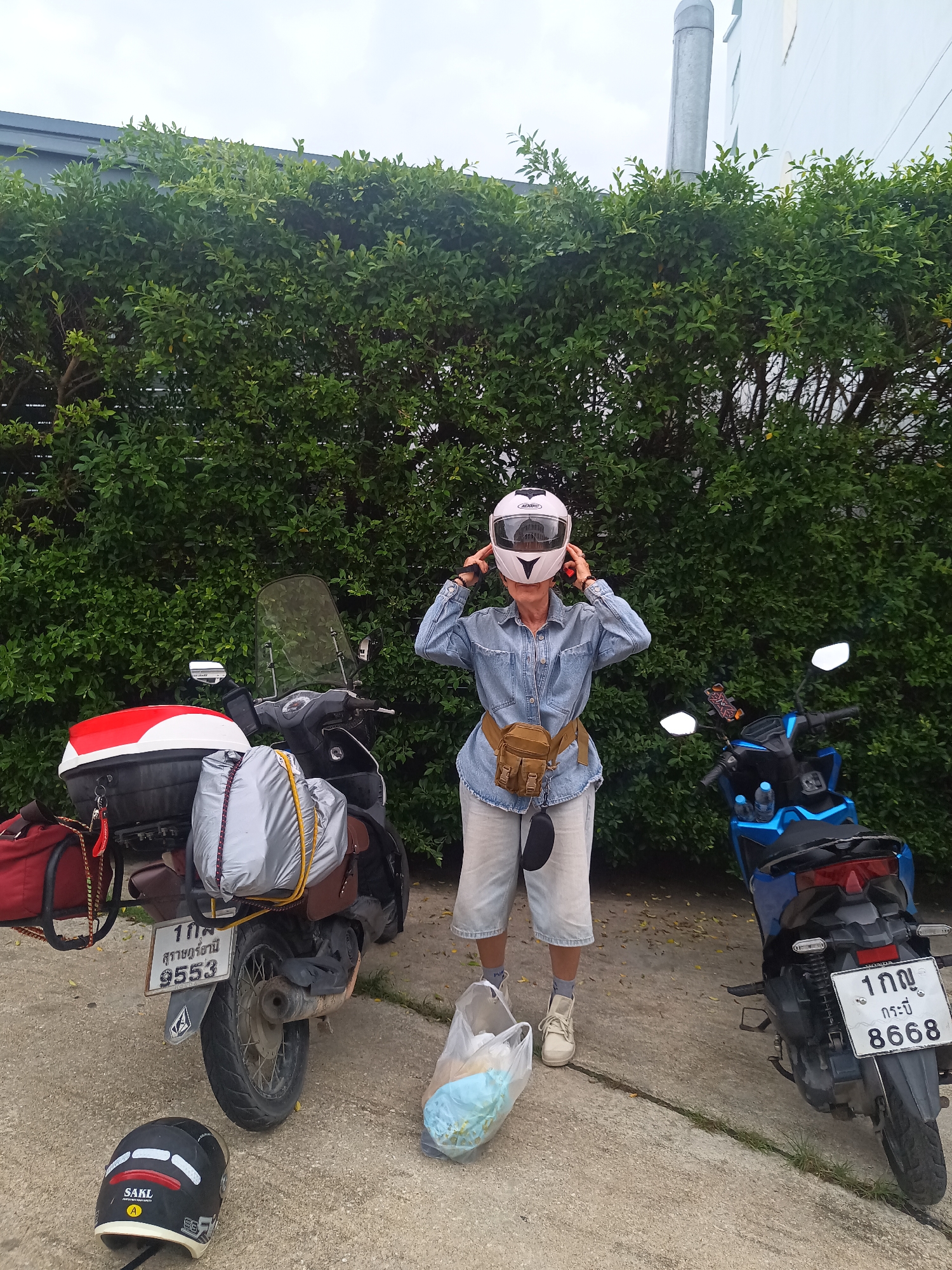 A person stands between two motorcycles outdoors in Ban Ao Nang, Thailand. The person is wearing a denim shirt, light-colored shorts, beige sneakers, a light pink hat, and a tan waist pack, and is adjusting a white motorcycle helmet. A black motorcycle helmet lies on the ground near the motorcycle on the left. The motorcycle on the left is black with a red and white storage box and silver luggage. The license plate on the left motorcycle reads "1 กค สุราษฎร์ธานี 9553". The motorcycle on the right is blue and black. The license plate on the right motorcycle reads "1 กญ กระบี่ 8668". A plastic bag filled with items sits on the ground near the person's feet. A tall green hedge forms the backdrop. A portion of a white building with a silver pipe is visible in the upper right. The ground is concrete. The sky is overcast.