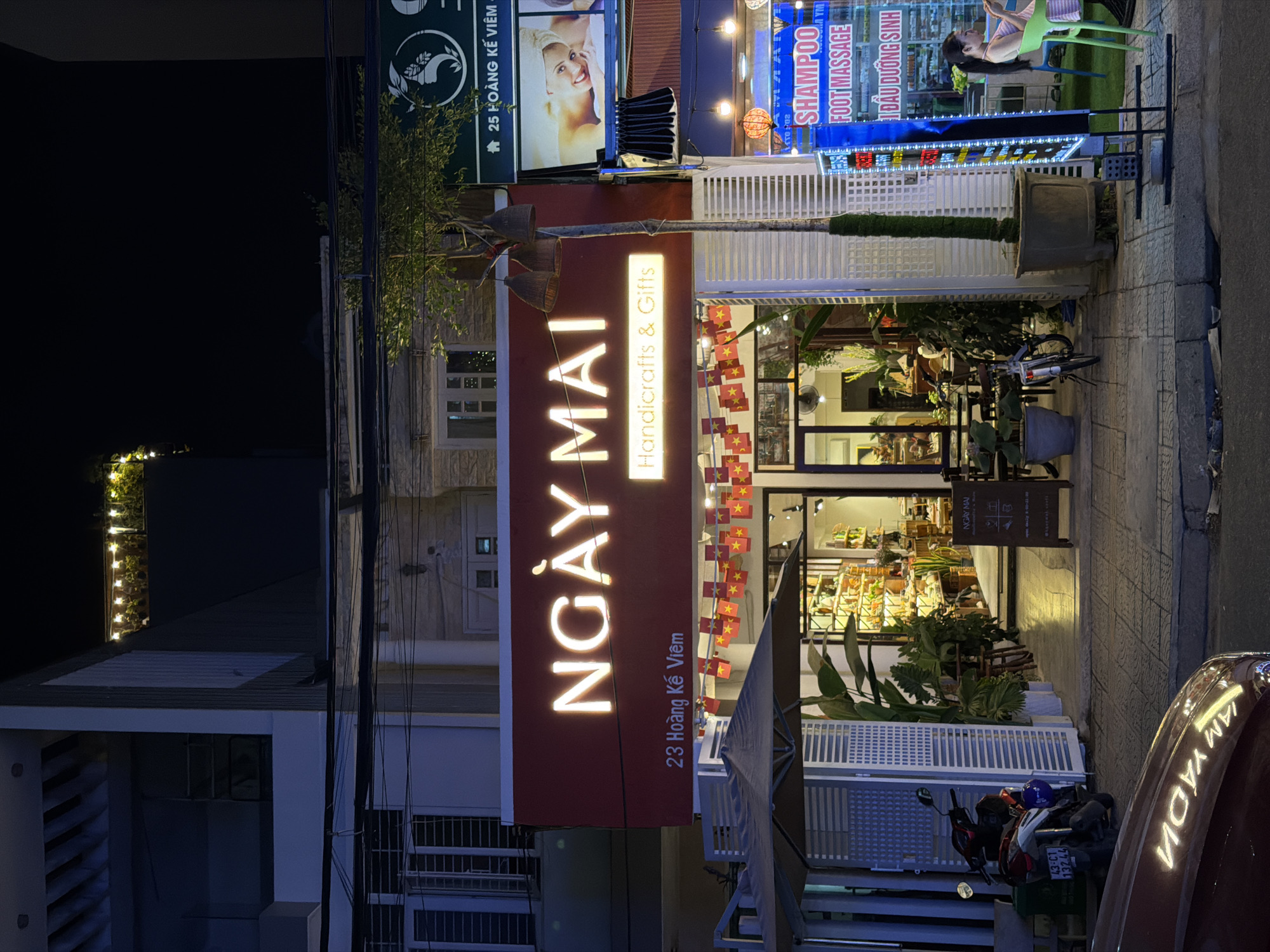 This is a nighttime exterior shot of a shop called "NGÀY MAI" (meaning "Tomorrow" in Vietnamese) located at 23 Hoàng Kế Viêm in Đà Nẵng, Vietnam. The shop appears to be a handicraft and gift store, as indicated by the signage. The shopfront is lit up, showcasing various items displayed inside, including what look like artisanal products and plants. In front of the shop, there are small Vietnamese flags decorating a white fence. A red car is parked on the right side of the image, reflecting the shop's lights. A motorcycle is parked in front of the shop on the left. Above and to the left of the main shop sign, there's another sign with an image of a woman and text that appears to be related to beauty services, mentioning "SHAMPOO" and "FOOT MASSAGE" in English and Vietnamese. The overall scene suggests a lively street in a Vietnamese city at night, with commercial activity and pedestrian traffic.