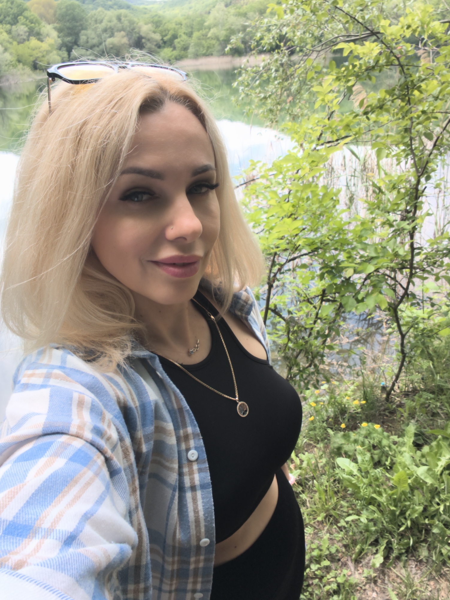 A fair-skinned woman with blonde hair and blue eyes poses for a selfie. She is wearing sunglasses on her head, a black top, a blue and white plaid shirt, and gold necklaces. She is standing in front of a body of water with trees and greenery in the background. The location is outdoors and appears to be near a lake or pond. Based on the prompt, the location is in the city of Терновский округ, Ukraine.