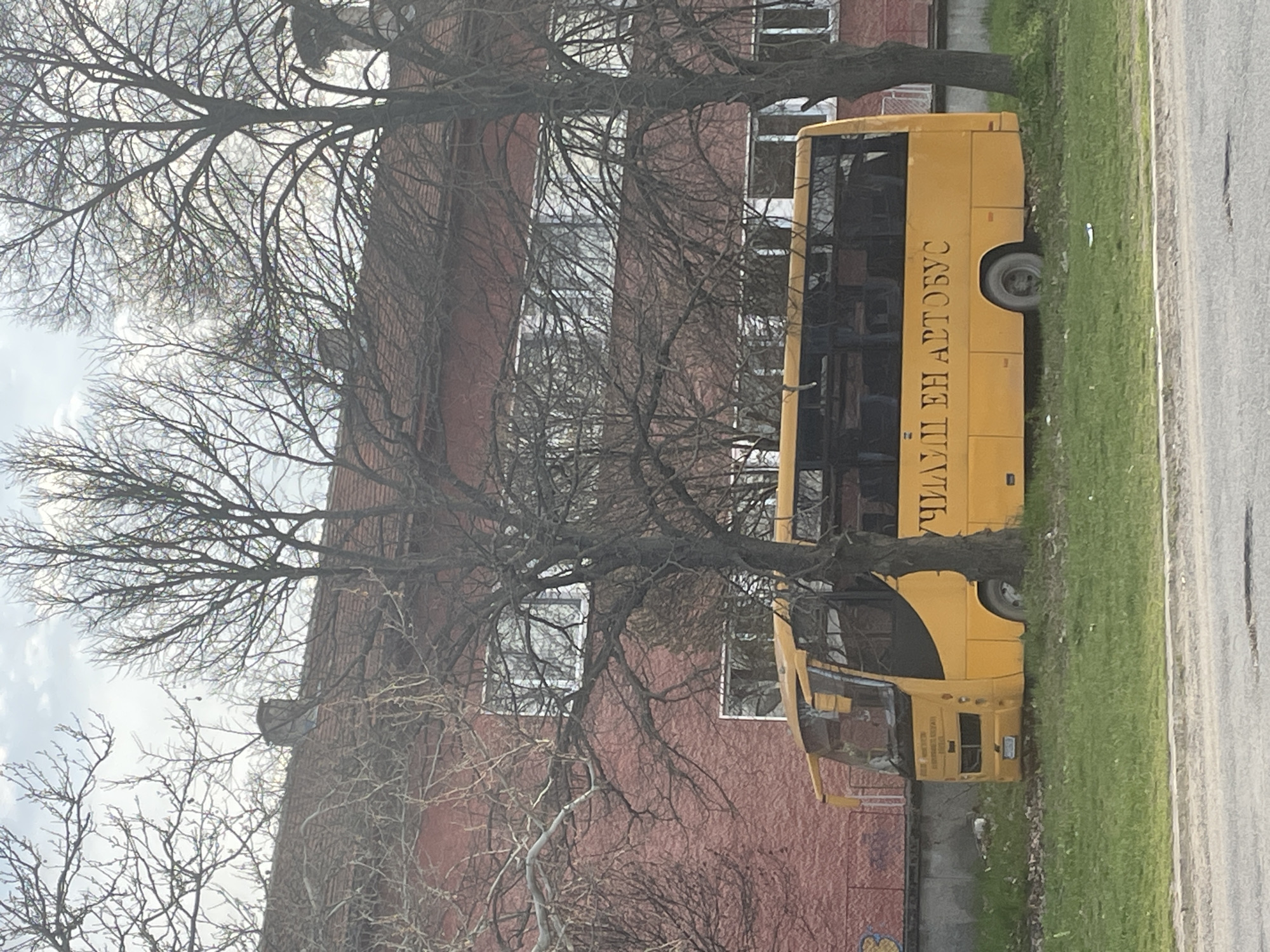 A yellow bus with Cyrillic text is parked on a grassy area alongside a street. The bus is positioned in front of a red building with multiple windows. Two bare trees are situated between the bus and the building, partially obscuring both. The scene is in Aprilovo, Bulgaria.