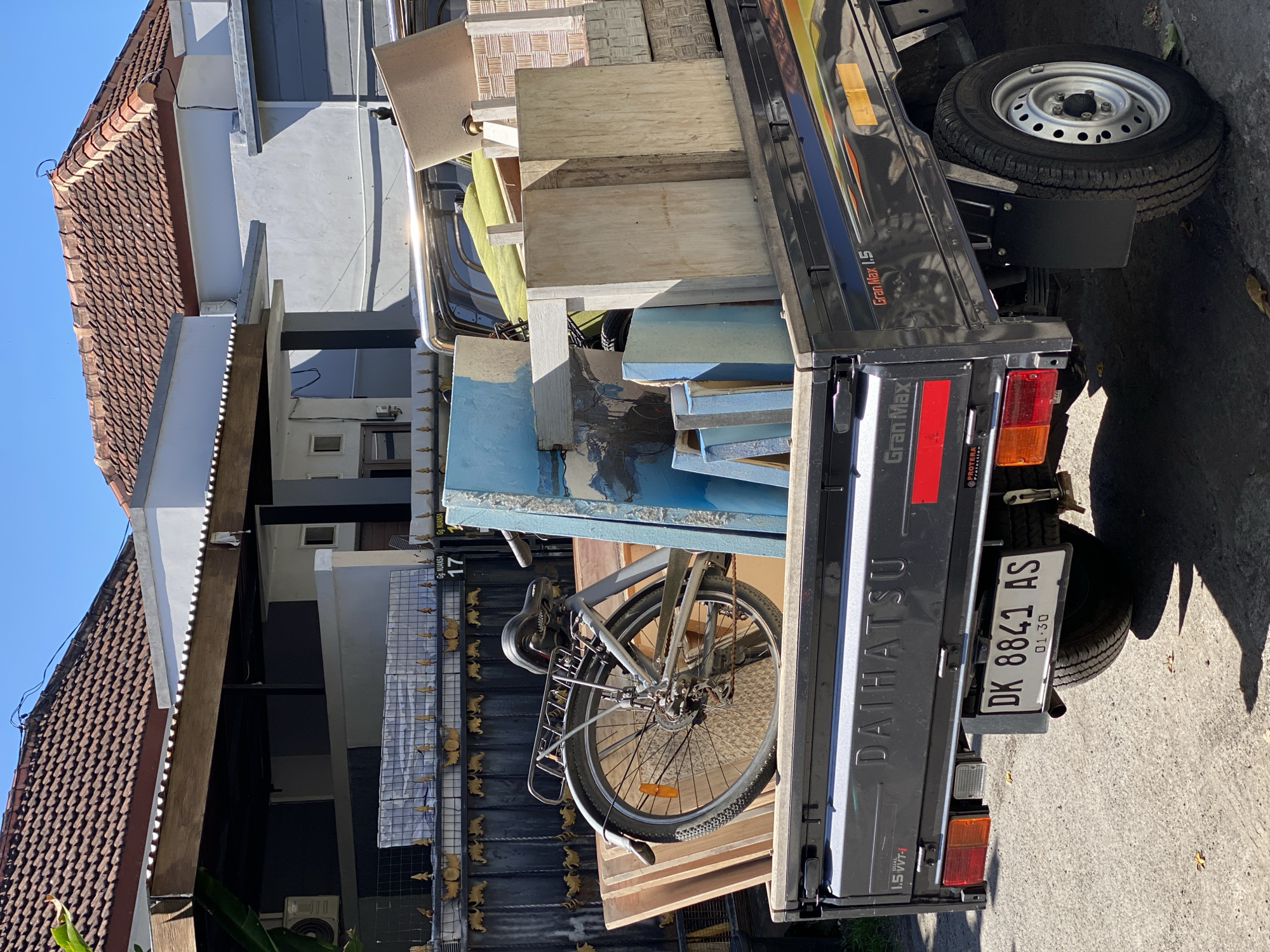 The image shows a flatbed Daihatsu Gran Max 1.5 pickup truck loaded with various items, including lumber, blue panels, a bicycle, and what appear to be furniture parts. The truck is parked on an asphalt surface, likely a street, in front of a residential area with houses featuring terracotta tiled roofs and fenced yards. The license plate of the truck is visible and reads "DK 8841 AS". Based on the license plate prefix "DK", the image is located in Bali, Indonesia. The presence of houses with distinct architectural styles, the style of the gate, and the license plate format suggest the location is within the city of Denpasar Barat, Indonesia. The scene is brightly lit by sunlight, indicating it is daytime. There are no people visible in the image.