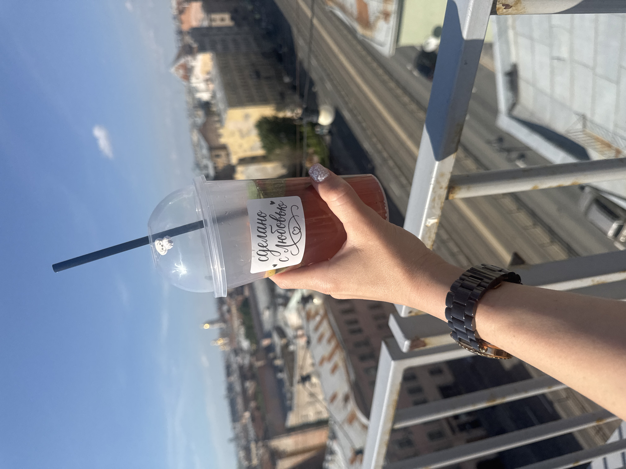 A person's arm holds a clear plastic cup filled with a red beverage and mint leaves. A black straw extends from the cup, and a small sticker of a white dog is visible on the lid. The cup has a label with Cyrillic text that reads "сделано с любовью," which translates to "made with love." The person wears a dark-colored, metallic watch on their wrist. The background shows a panoramic view of Saint Petersburg, Russia, with distinct architecture and buildings, including what appears to be a dome of a church in the distance. The scene suggests an elevated viewpoint, possibly from a rooftop or a high balcony overlooking the city under a clear blue sky.
