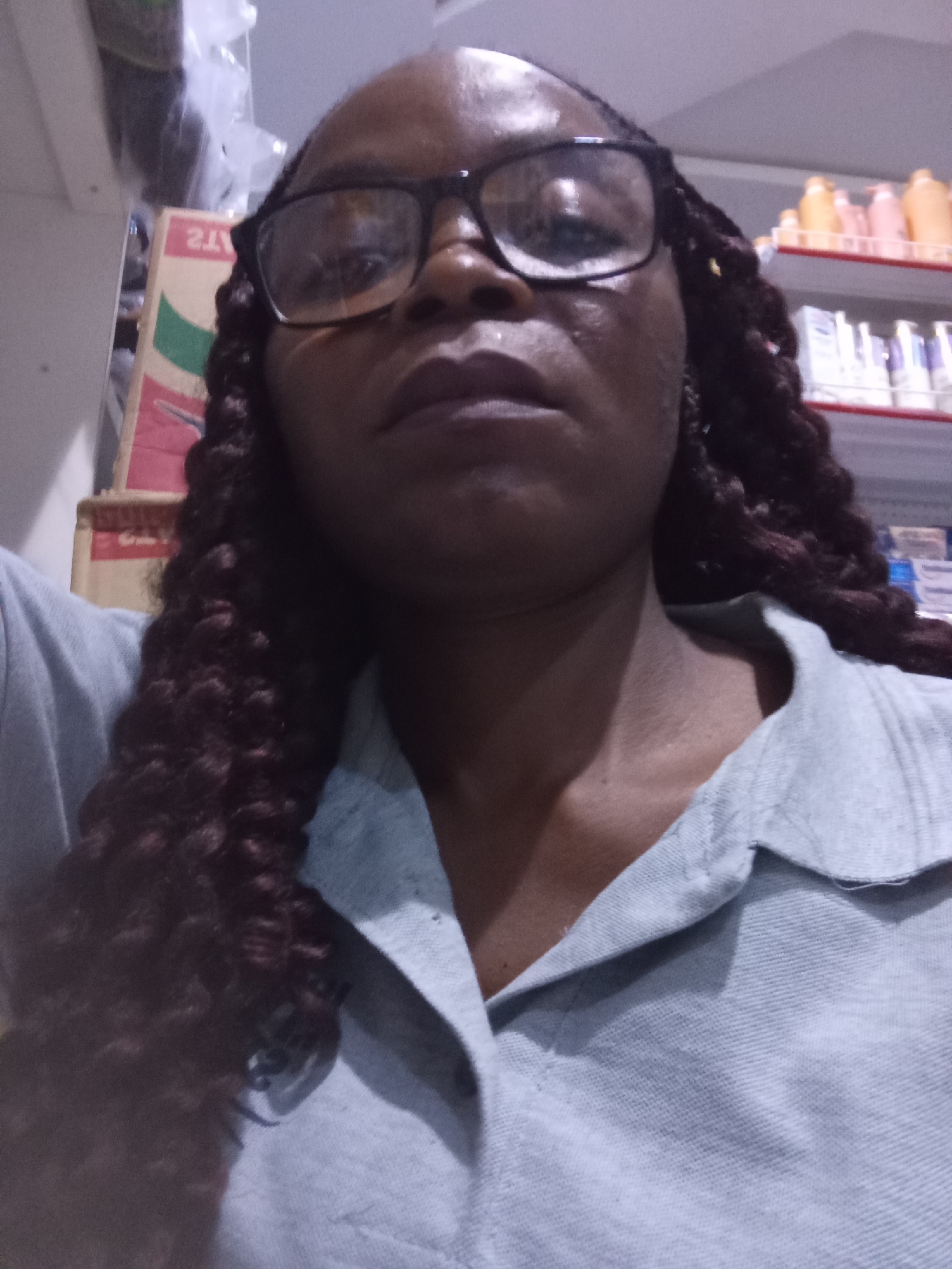 The image shows a close-up, selfie-style shot of a Black woman from the chest up. She is wearing black-rimmed glasses and a light blue collared shirt. Her hair is styled in dark, braided extensions that fall over her shoulders. The background appears to be a store or market setting, with shelves stocked with various products, including what seem to be bottles of shampoo or lotion, and boxes. The lighting is artificial and somewhat dim, casting a slight shadow on her face. The overall impression is that of a casual snapshot taken indoors.