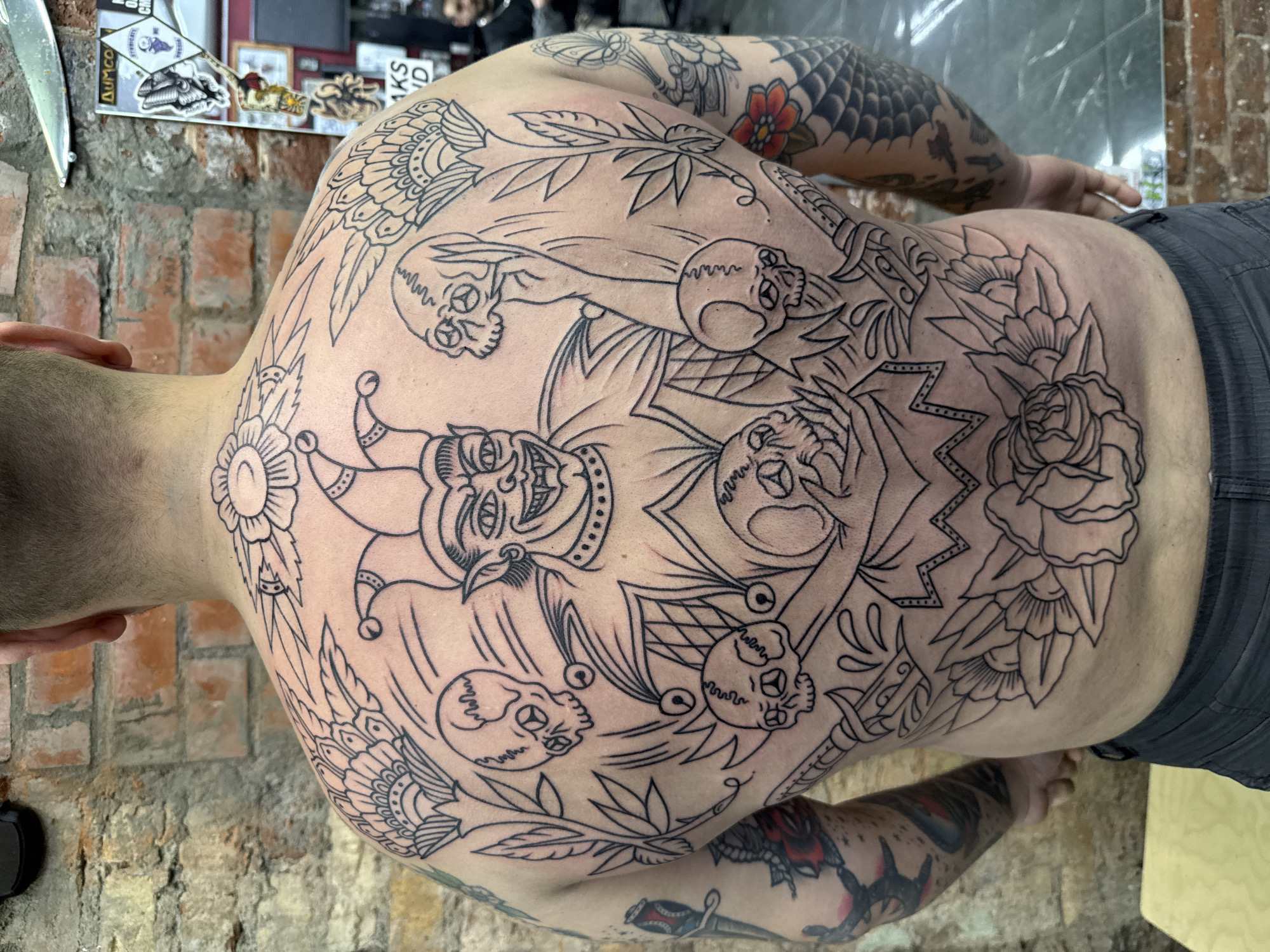The image shows a man's back and shoulders, extensively tattooed with a detailed black ink design. The tattoo features multiple elements including a central jester figure, skulls, flowers (roses and possibly sunflowers), and decorative foliage. Additional tattoos are visible on his arms. The location appears to be a tattoo studio, based on the brick wall and what looks like tattoo-related stickers and equipment visible in the background. The man is lying down, presumably receiving the tattoo. The image does not have information related to the city of Moscow, Russia.