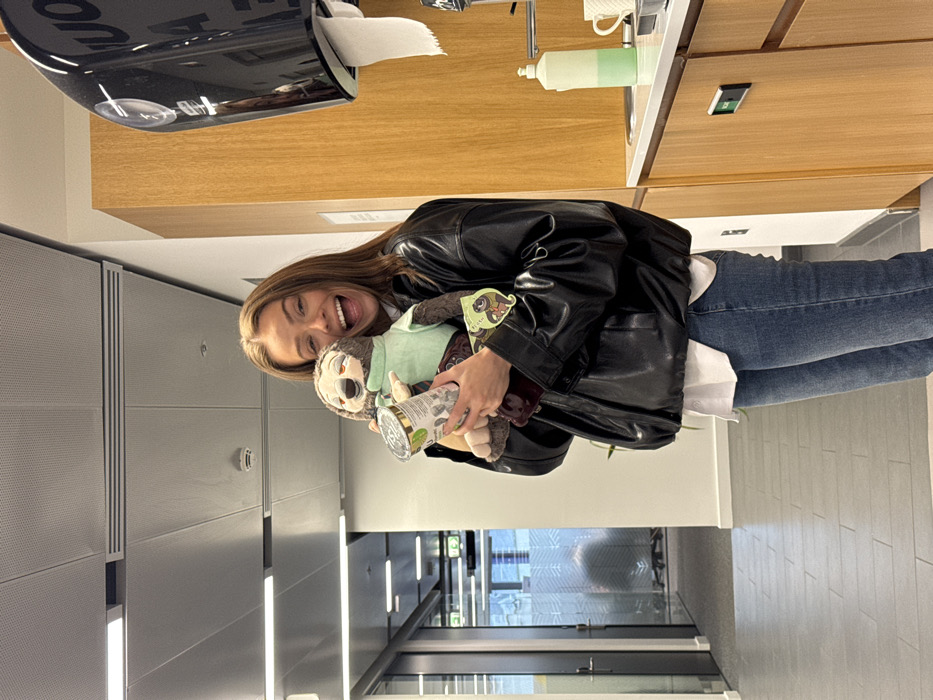 A female subject, appearing to be in her 20s or early 30s with medium brown hair, stands smiling broadly at the camera. She wears a black bomber-style jacket over a white shirt and blue jeans. She holds a brown sloth plush toy, wearing a light green shirt, in her left arm, and a cylindrical white and green can-like object in her right hand. A small tag with a cartoon sloth is attached to the plush toy.
The background on the left reveals a modern office corridor with grey tiled flooring, white walls, and a suspended ceiling featuring integrated lighting and a visible smoke detector. A glass-partitioned office space with interior furniture is visible further down the corridor. The background on the right features a kitchenette area with light wooden cabinetry, a white countertop, a black wall-mounted paper towel dispenser, a light green two-tone liquid bottle, and a white mug. The architecture and furnishing suggest a contemporary commercial building or office environment within the city of London, United Kingdom.