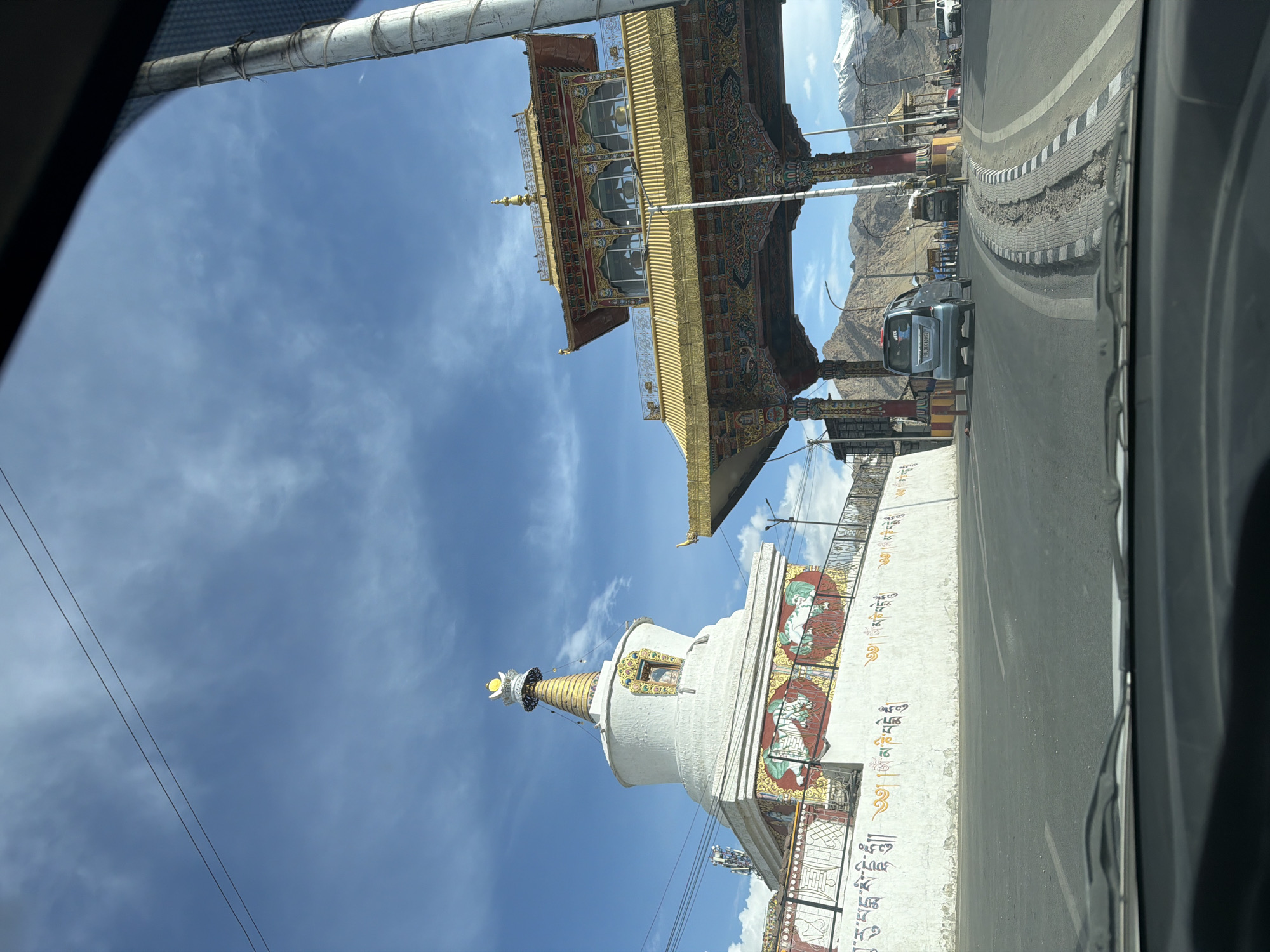 The image captures a view from inside a vehicle on a road in Leh, India, with a clear blue sky and scattered clouds overhead. On the left, a white stupa with golden accents and intricate murals is visible. To the right, a large, ornate golden gate with colorful Tibetan architectural details spans the road. Beyond the gate, a convoy of vehicles is seen approaching, with mountainous terrain and snow-capped peaks forming the backdrop. The road itself has a dividing strip with white and black markings, and some areas show signs of wear and tear. A utility pole stands tall on the right side of the frame. The scene suggests a journey into or out of the city, with prominent religious and cultural landmarks marking the entrance.